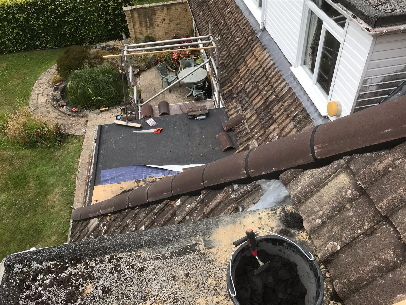 Overhead view of a house roof under construction, with new flat roof membrane, terracotta roof tiles, and a bucket of mortar on the roof.