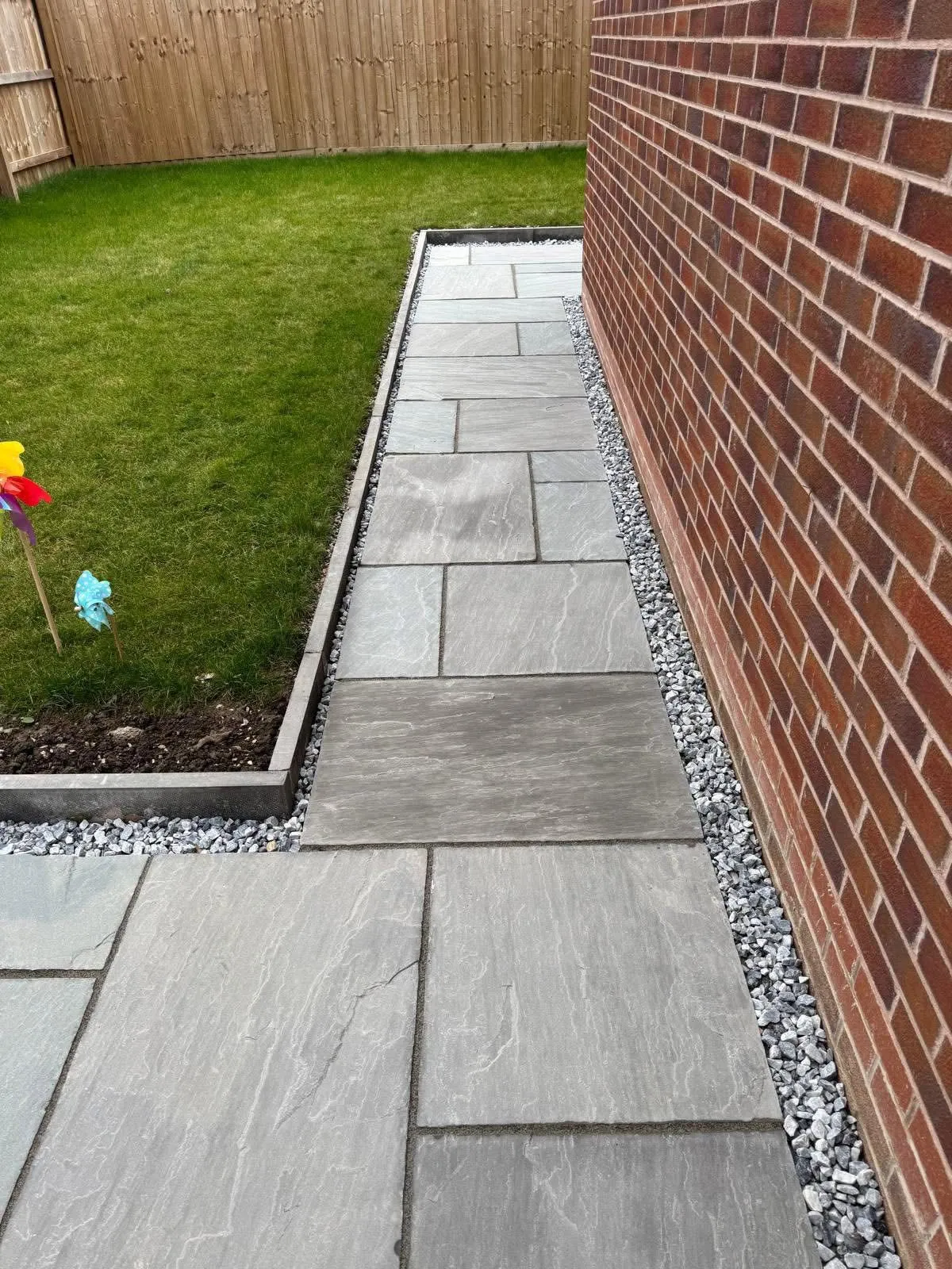 Grey Indian sandstone slab pathway alongside a brick house wall in a residential garden
