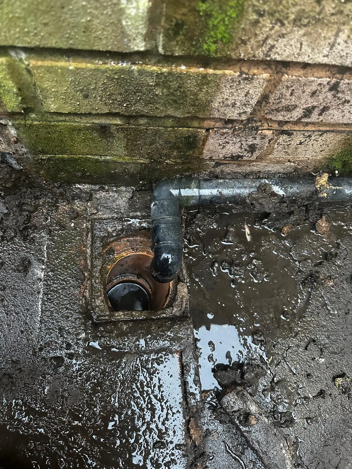 Blocked external drain or gully with dirty, standing water and mud next to a house wall.