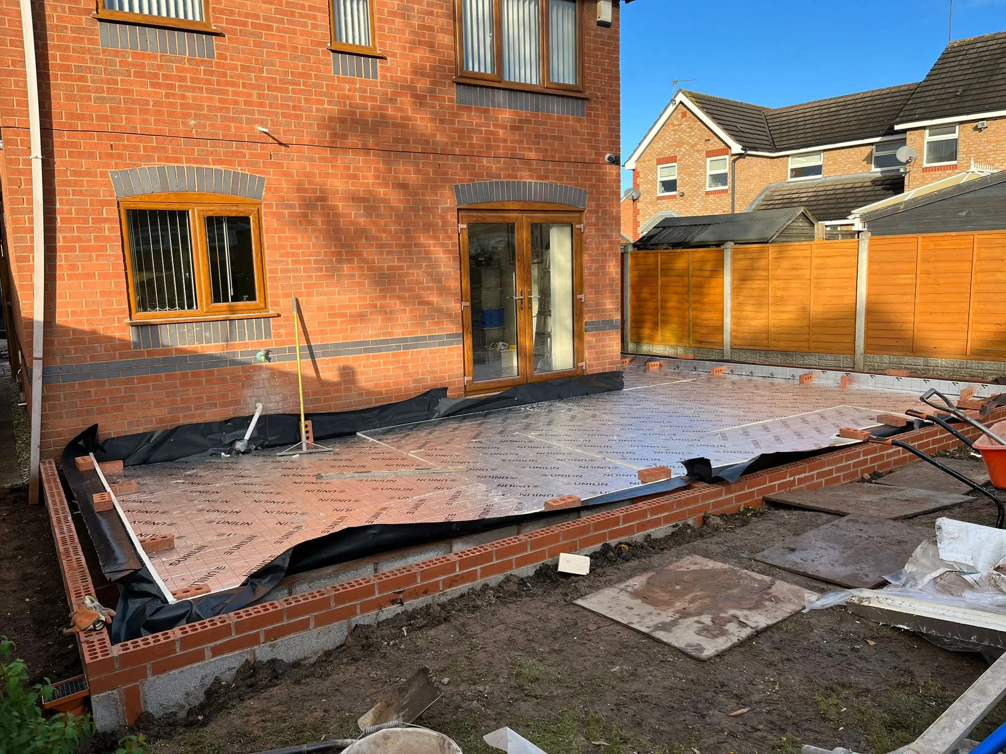 A home extension under construction with new brick foundations, partial walls, and insulation laid over the ground, viewed from the garden.