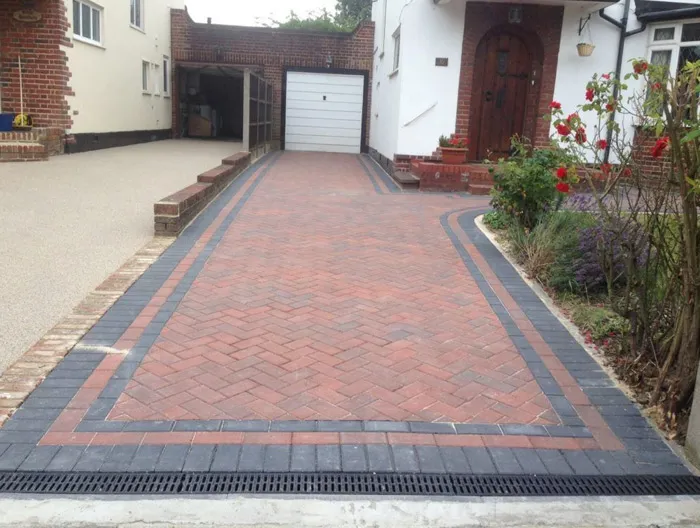 Newly laid block paving driveway with a red herringbone pattern, dark grey border, drainage channel, and an adjacent light-coloured resin-bound driveway leading to a garage.
