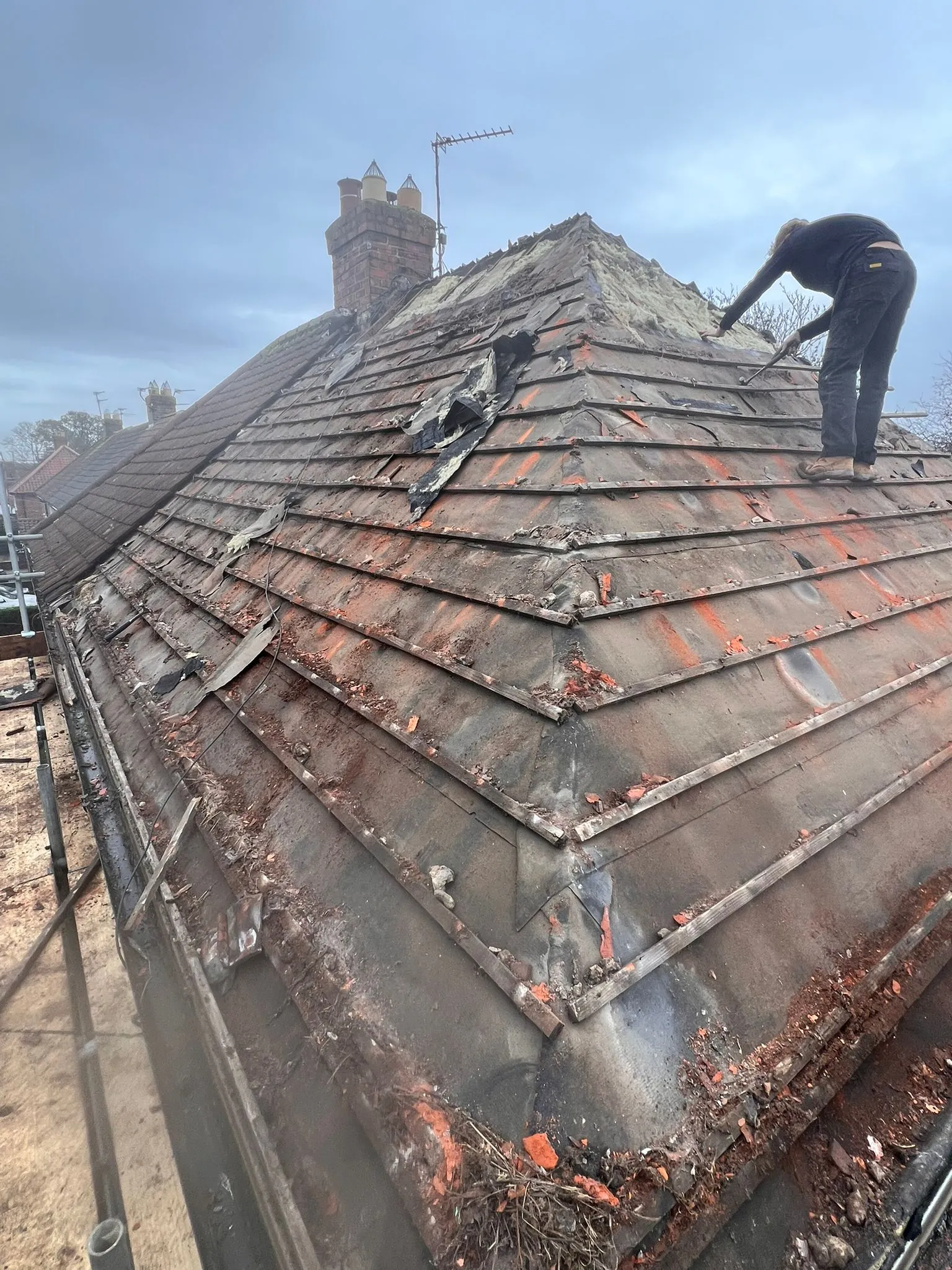 A roofer on scaffolding working on a very old and heavily damaged pitched roof with many broken tiles and debris, undergoing replacement or major repair.
