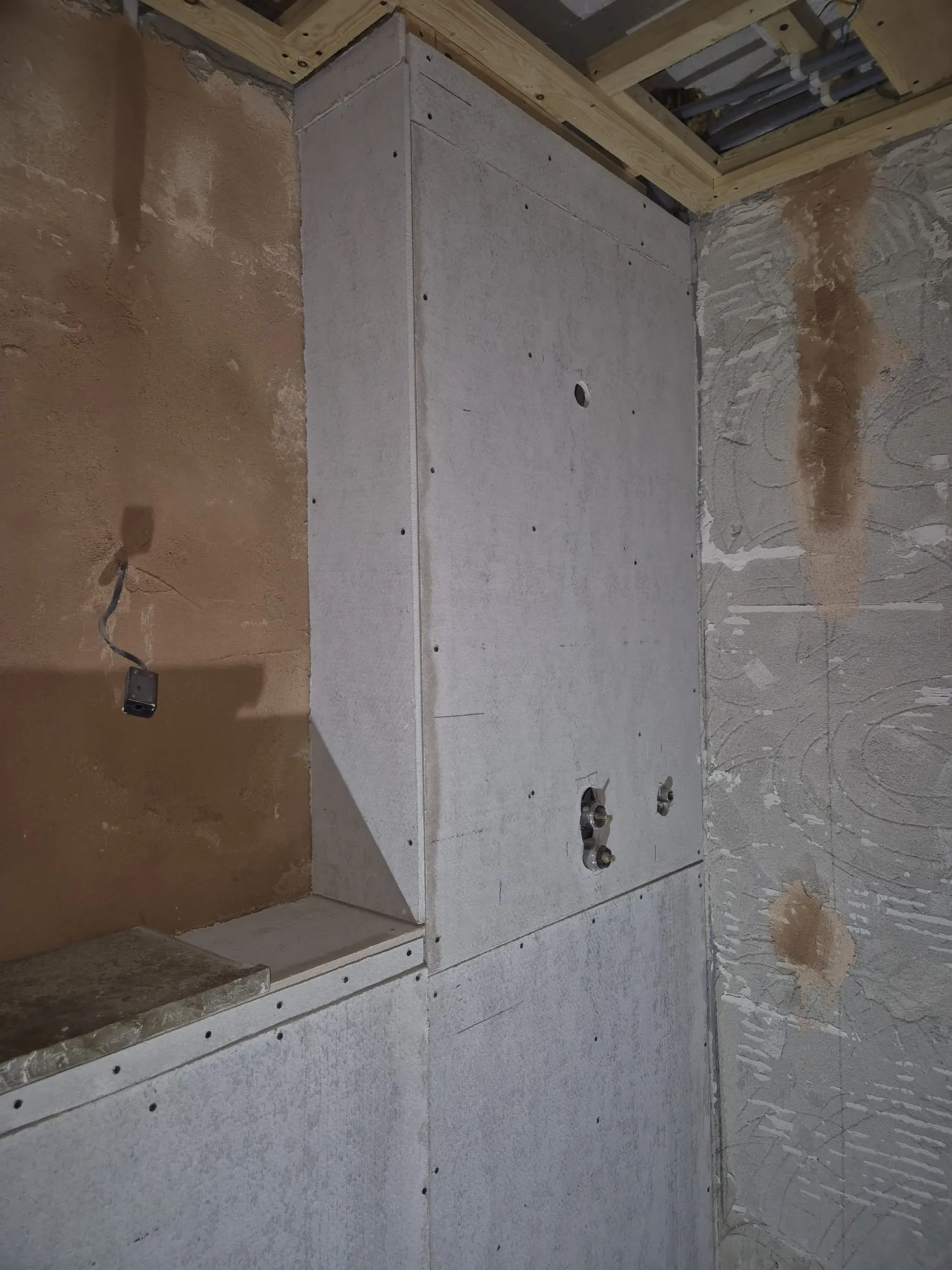 An unfinished room during refurbishment, showing grey plasterboard walls with exposed plumbing fittings for a mixer tap, electrical wiring, and a raw plastered wall section. Wooden joists are visible on the ceiling.