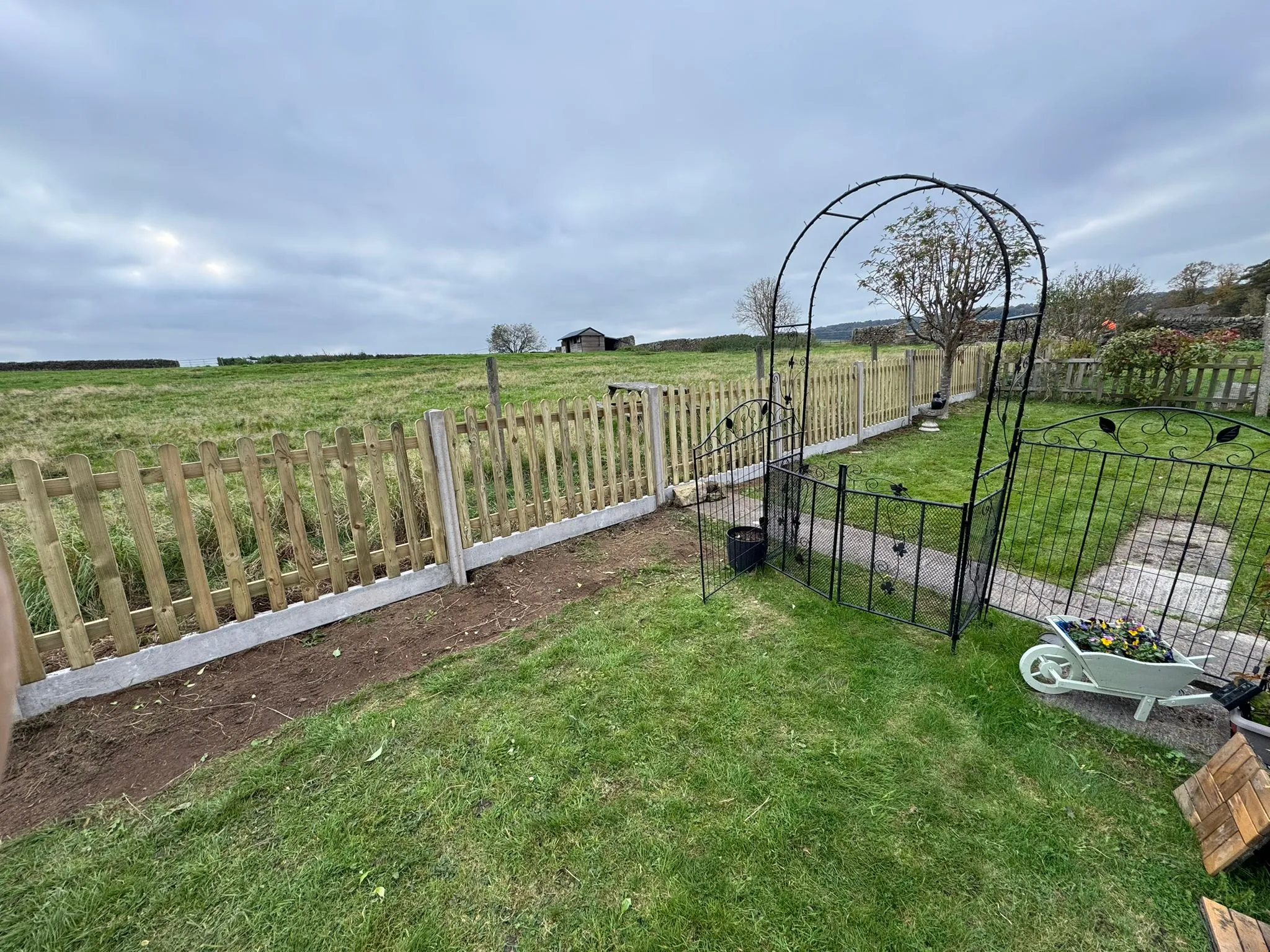 Newly installed wooden picket fence with concrete posts and gravel boards, a metal garden arch, and a green lawn, bordering a field.