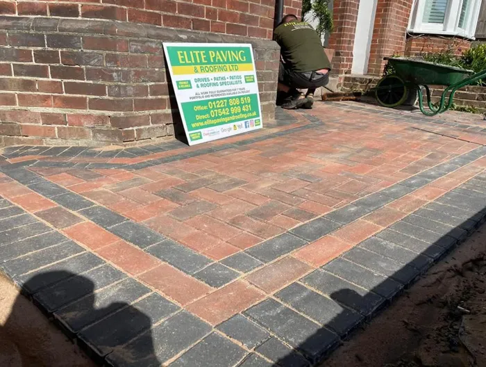 Newly laid block paving with red and grey bricks in a herringbone pattern, featuring a dark border, next to a brick wall with a company sign and a worker.