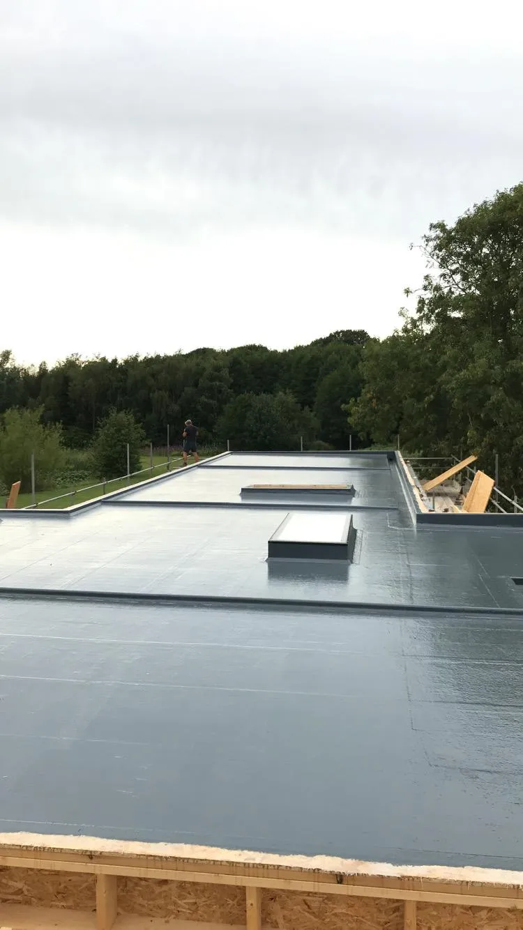 A completed modern grey flat roof installation with several integrated rooflights, showing a roofer in the distance against a backdrop of trees and an overcast sky.