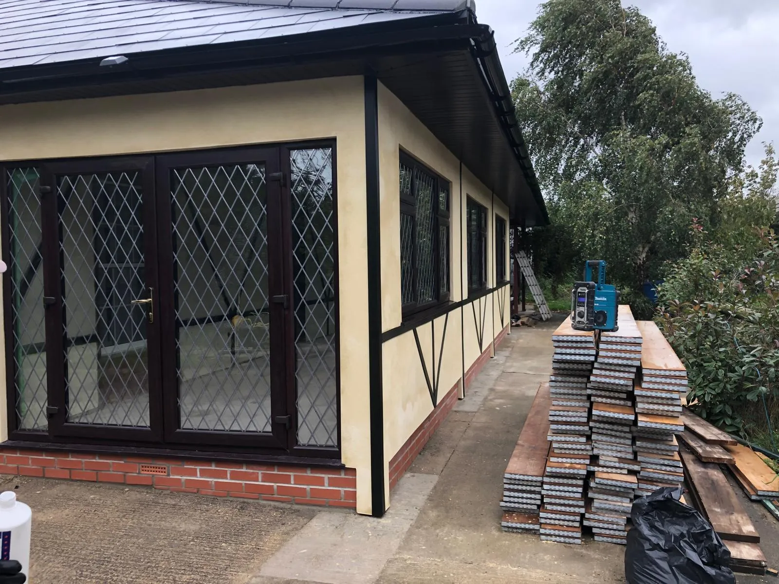 Exterior of a newly renovated house or extension featuring rendered walls, timber-effect accents, dark brown French doors with diamond leaded glass, new windows, and a dark grey roof with matching fascias and guttering. Building materials are stacked outside.