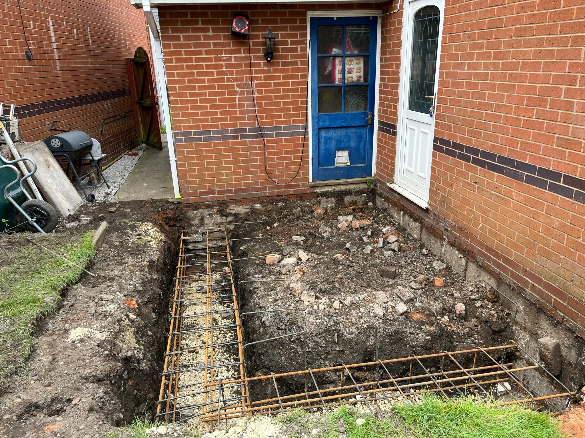 Foundations being laid with steel rebar for a new home extension next to a brick house.
