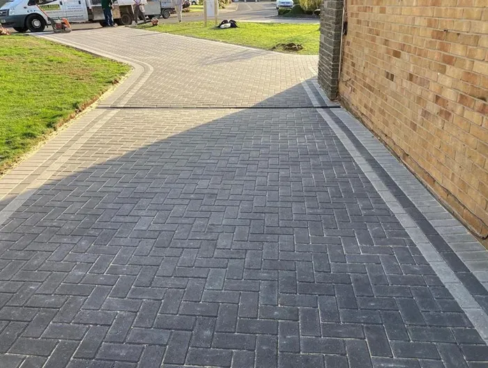 Newly laid dark grey block paved driveway with light border and Aco drainage channel, connecting to a lighter block paved path on a sunny day.