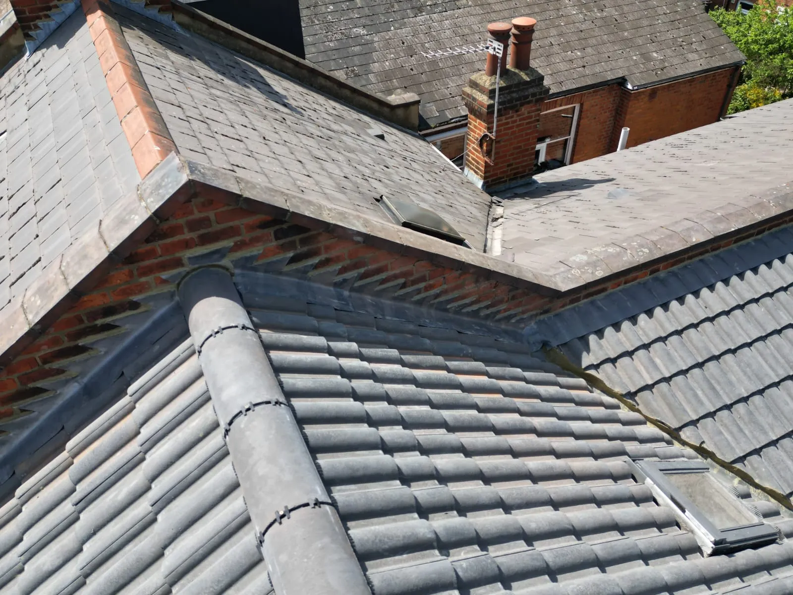 An elevated view of a residential roof showing modern grey tiles, a skylight, lead flashing along a brick parapet, and an older tiled roof with a chimney stack in the distance, illustrating diverse roofing work.