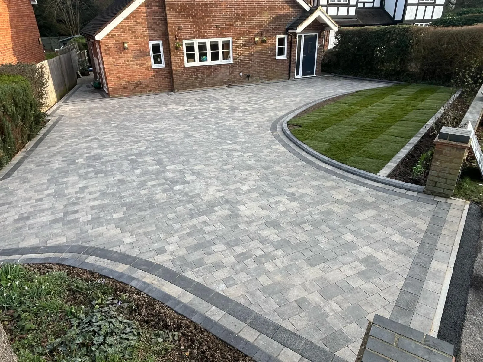 Newly installed grey block paving driveway with dark block edging and a new turf lawn in front of a red brick house.