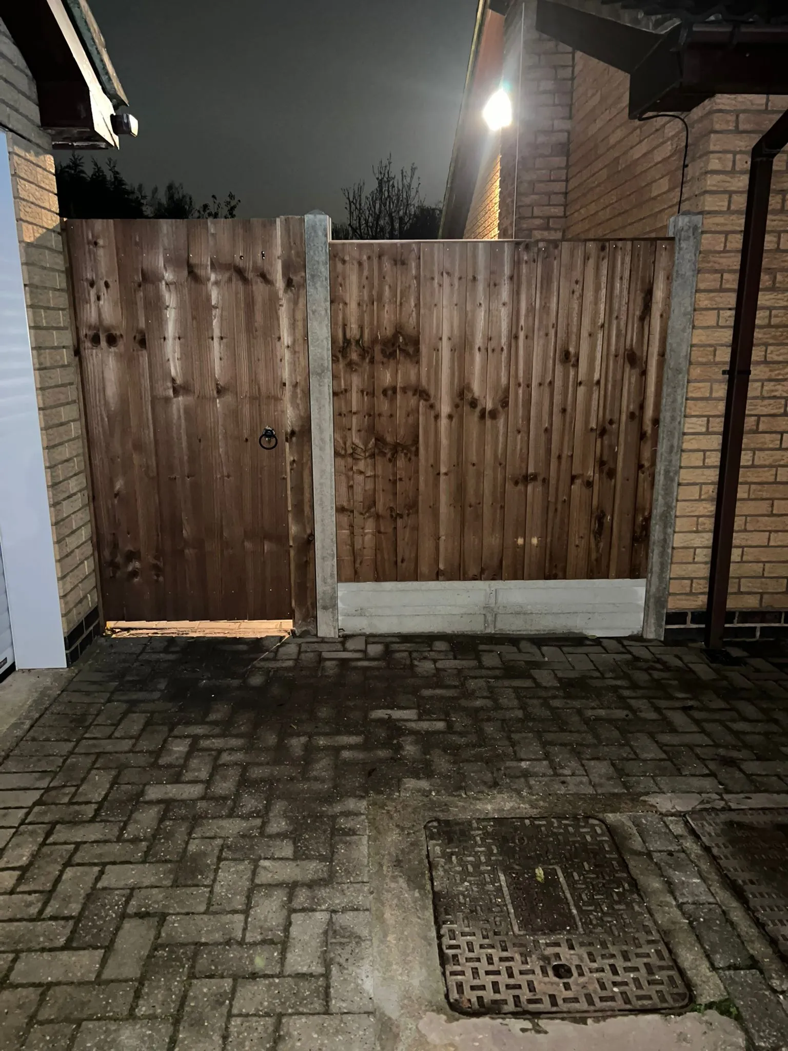 A wooden garden gate and fence with block paving, concrete posts, and a drain cover under artificial light at night.