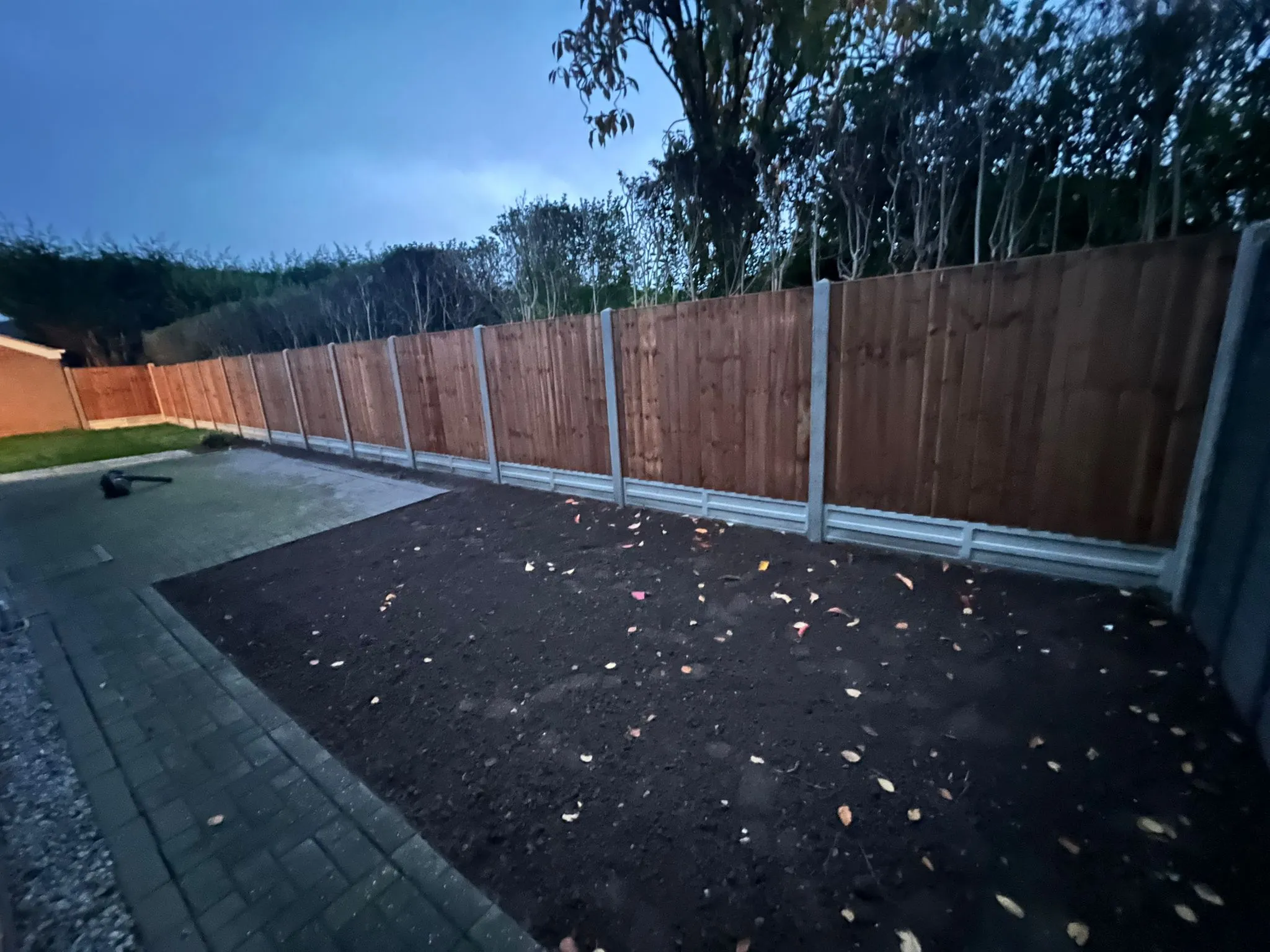 New wooden closeboard garden fence with concrete posts, prepared dark soil for landscaping, and paved pathway at twilight.