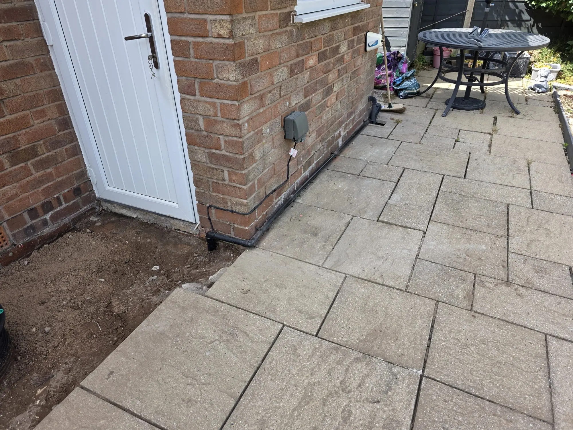 Exposed soil and pipework next to a brick house after patio slabs were removed for drain repairs.