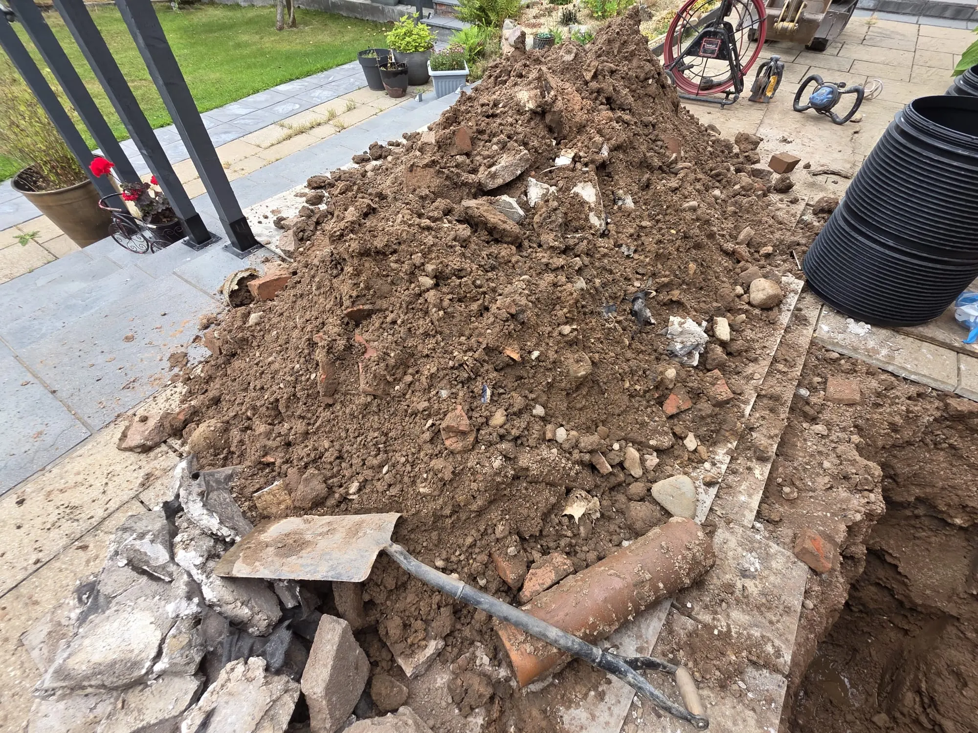 A drain excavation site showing a pile of soil, a trench, and drainage repair equipment on a patio