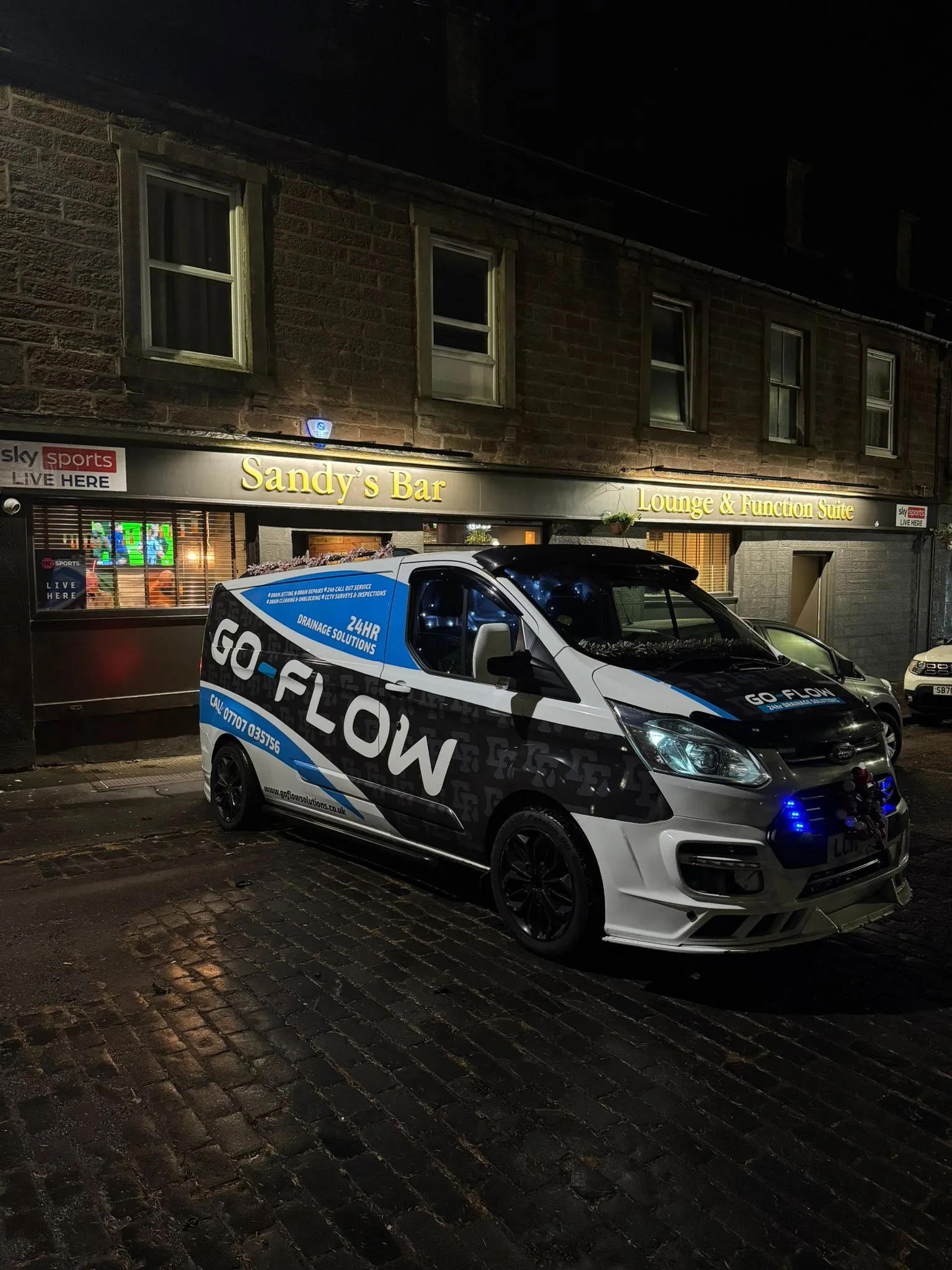 A Go Flow Solutions branded van with '24HR Drainage Solutions' on the side, parked at night outside a building with 'Sandy's Bar'.