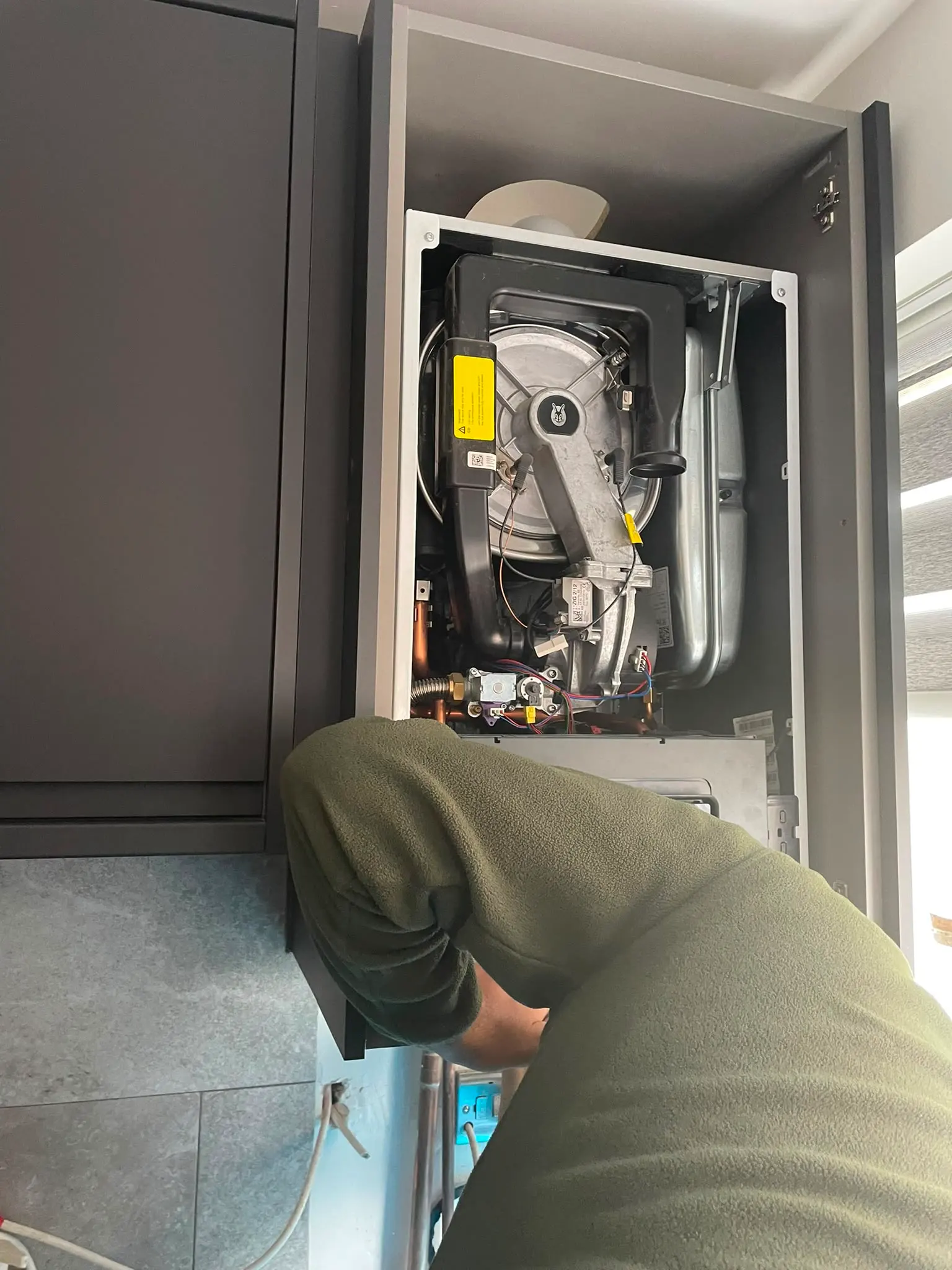 A plumber repairing or servicing a hot water boiler inside a kitchen cabinet