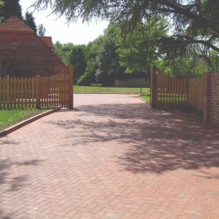 A wide block paved driveway entrance with open wooden gates on either side, a wooden building to the left, and trees in the background.