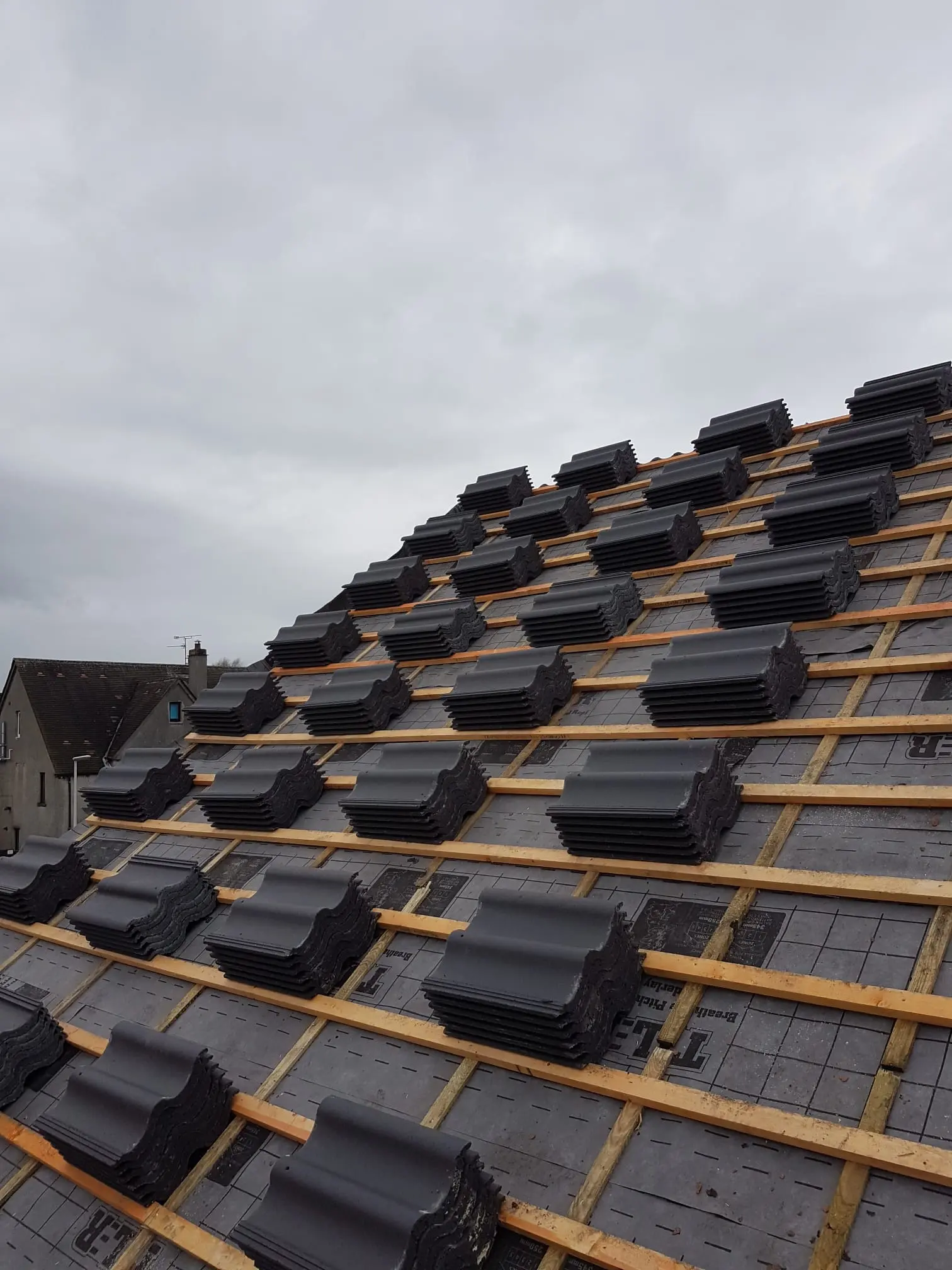 A picture of a new tiled roof installation in progress showing membrane, wooden battens, and stacks of grey roof tiles ready to be laid.