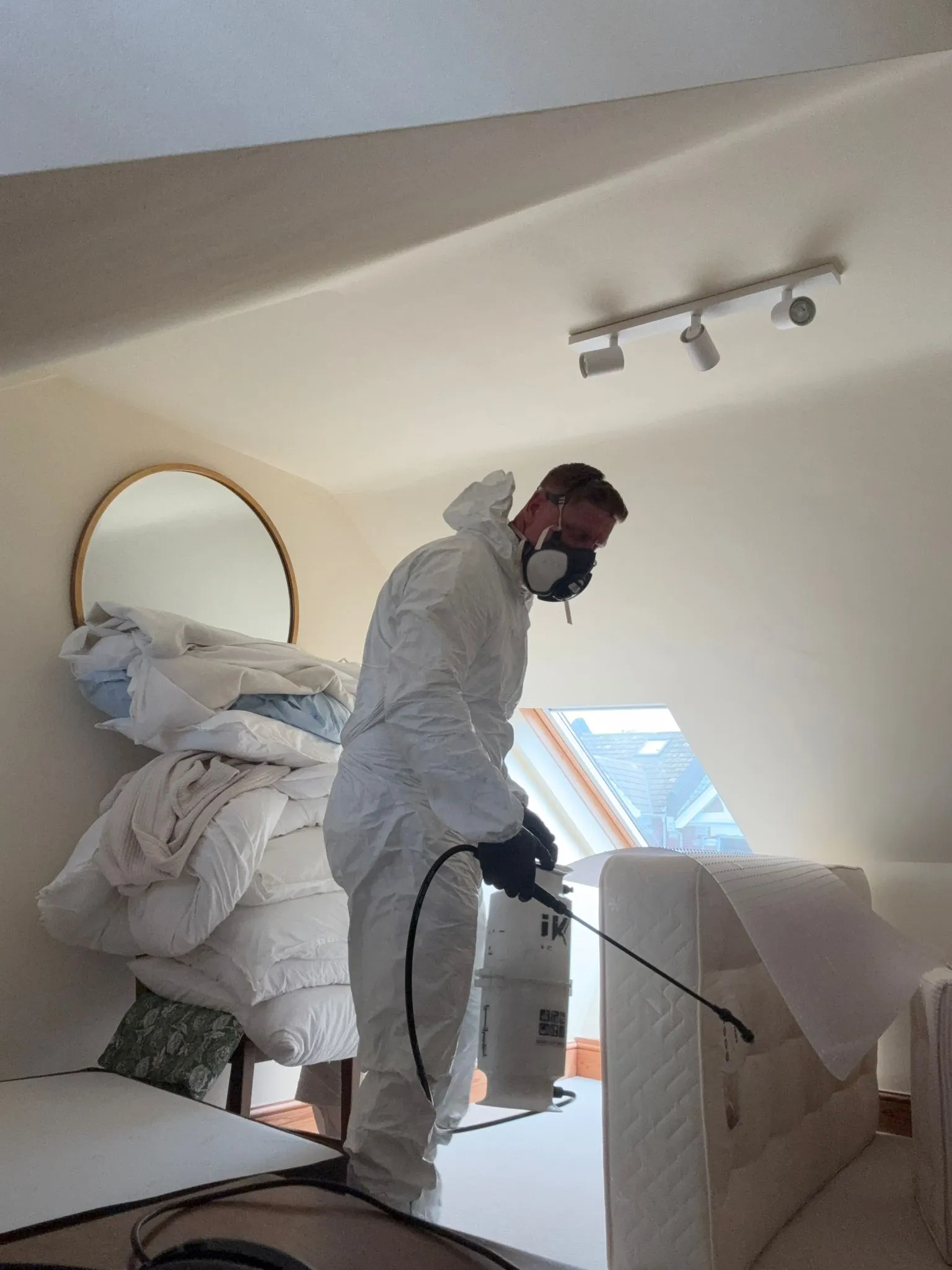 A pest control technician in a white protective suit and mask spraying a mattress for bed bug treatment.
