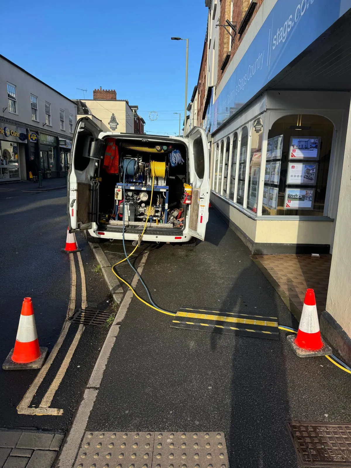 A white drainage service van with jetting equipment and hoses on a street, surrounded by safety cones, ready for drain unblocking.