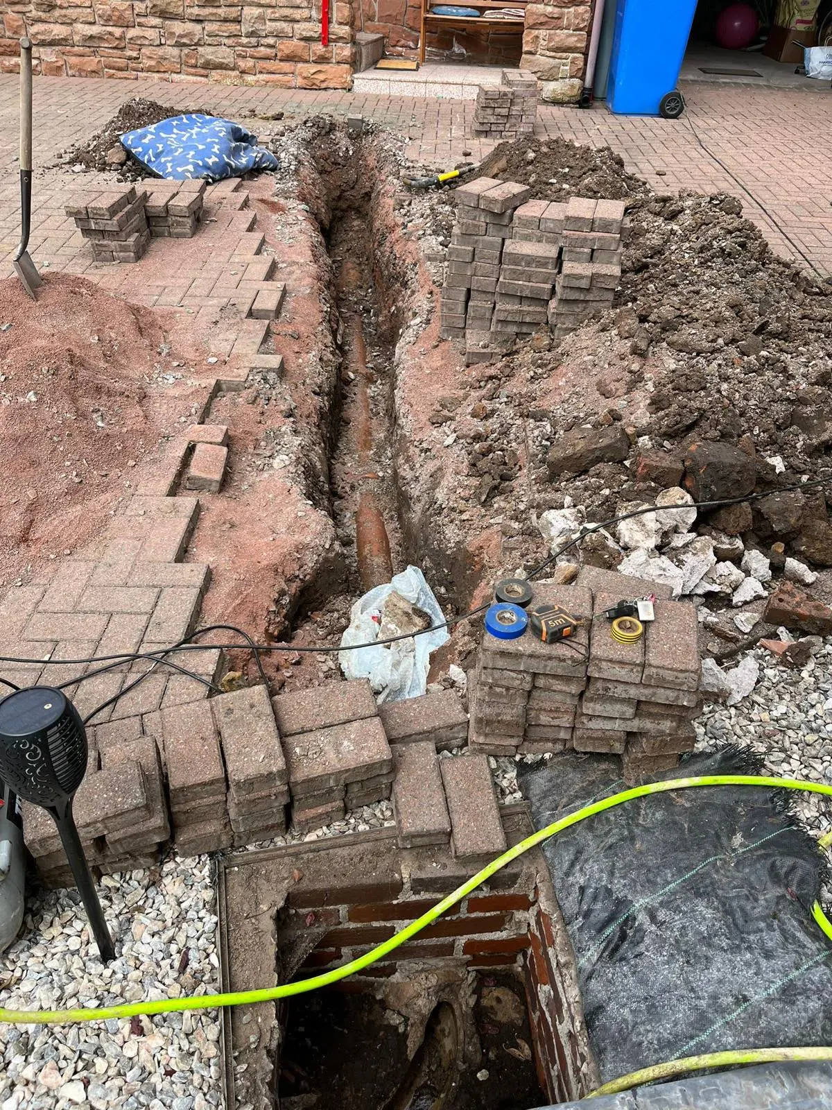 A drainage excavation trench on a block-paved driveway revealing an underground pipe for replacement, alongside an open brick manhole chamber.