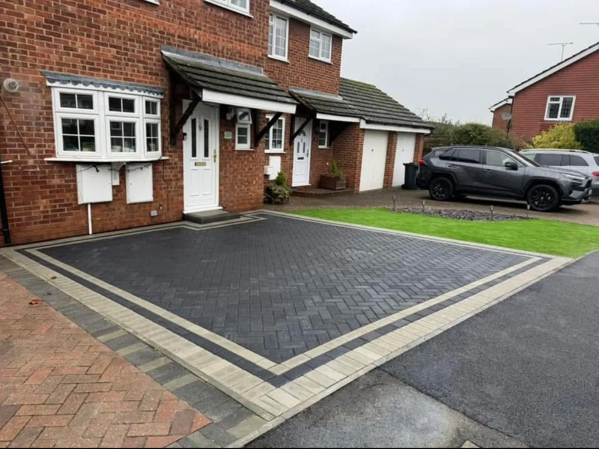 A newly installed charcoal block paved driveway with a light grey border in front of a brick house.