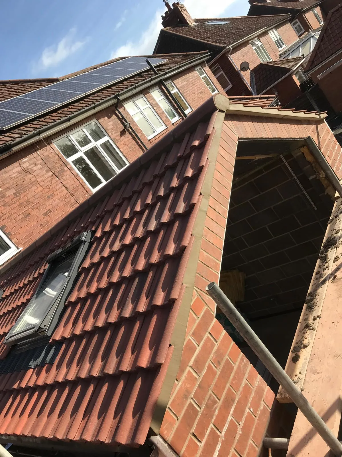 A close-up view of a newly tiled pitched roof section on a brick building, featuring a newly installed Velux-style roof window. Scaffolding is visible, and in the background, another house with solar panels on its roof can be seen.