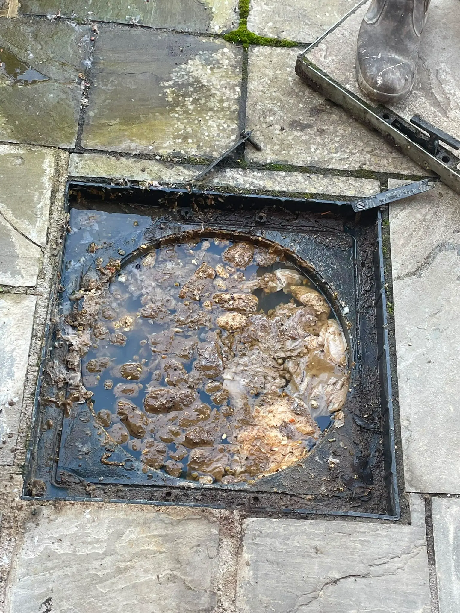 A heavily blocked outdoor sewer drain filled with waste and water with the cover removed
