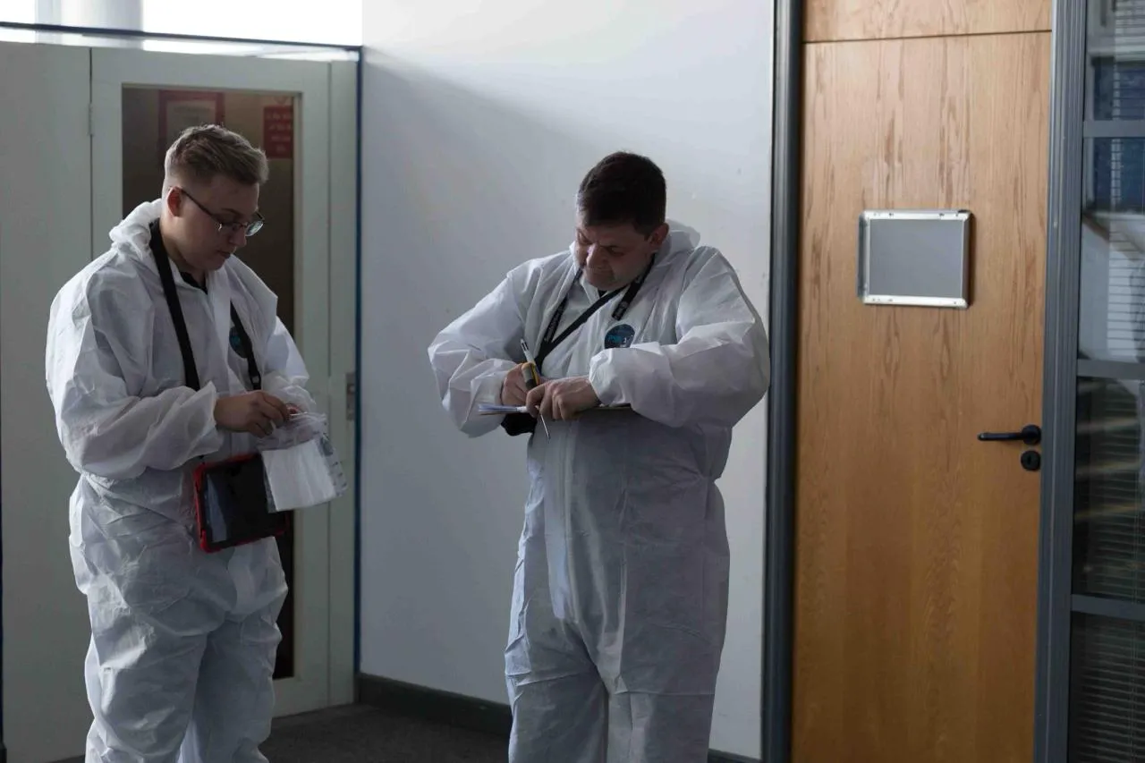 Two asbestos surveyors in full PPE, one holding a sample kit and the other writing on a clipboard, inside a modern building.