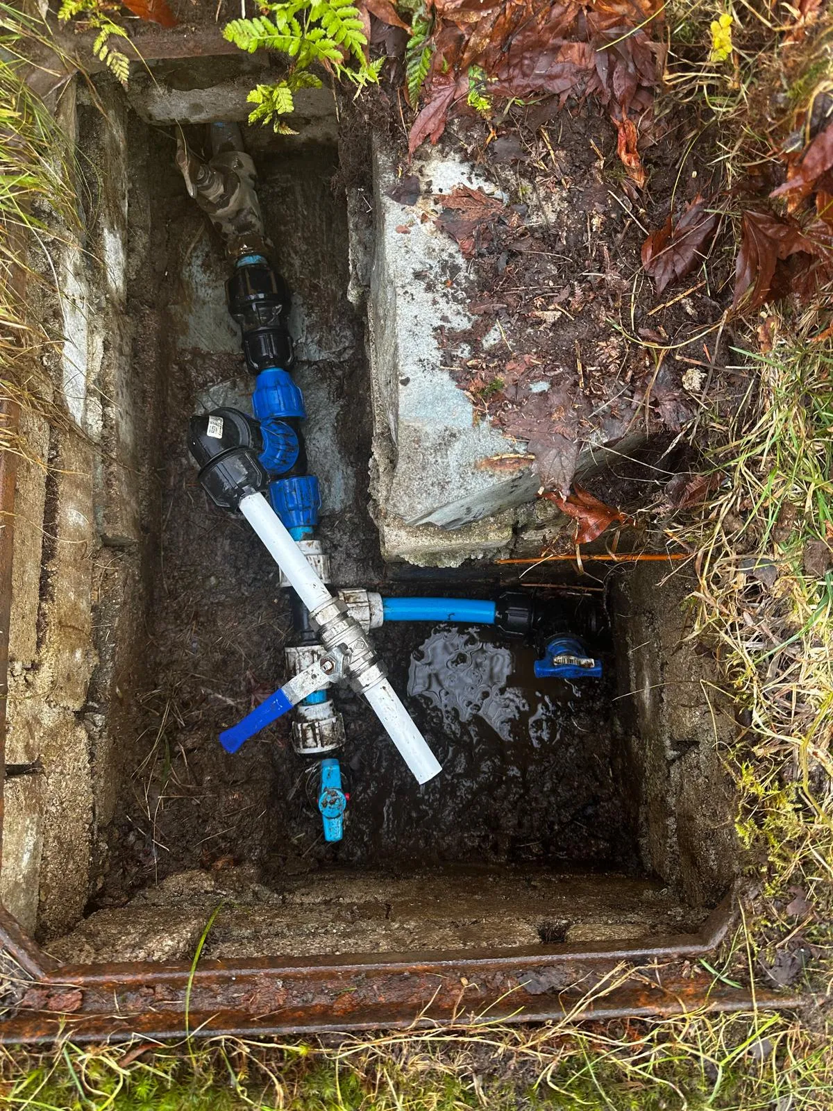 An outdoor utility access pit showing water pipes and valves with muddy standing water.