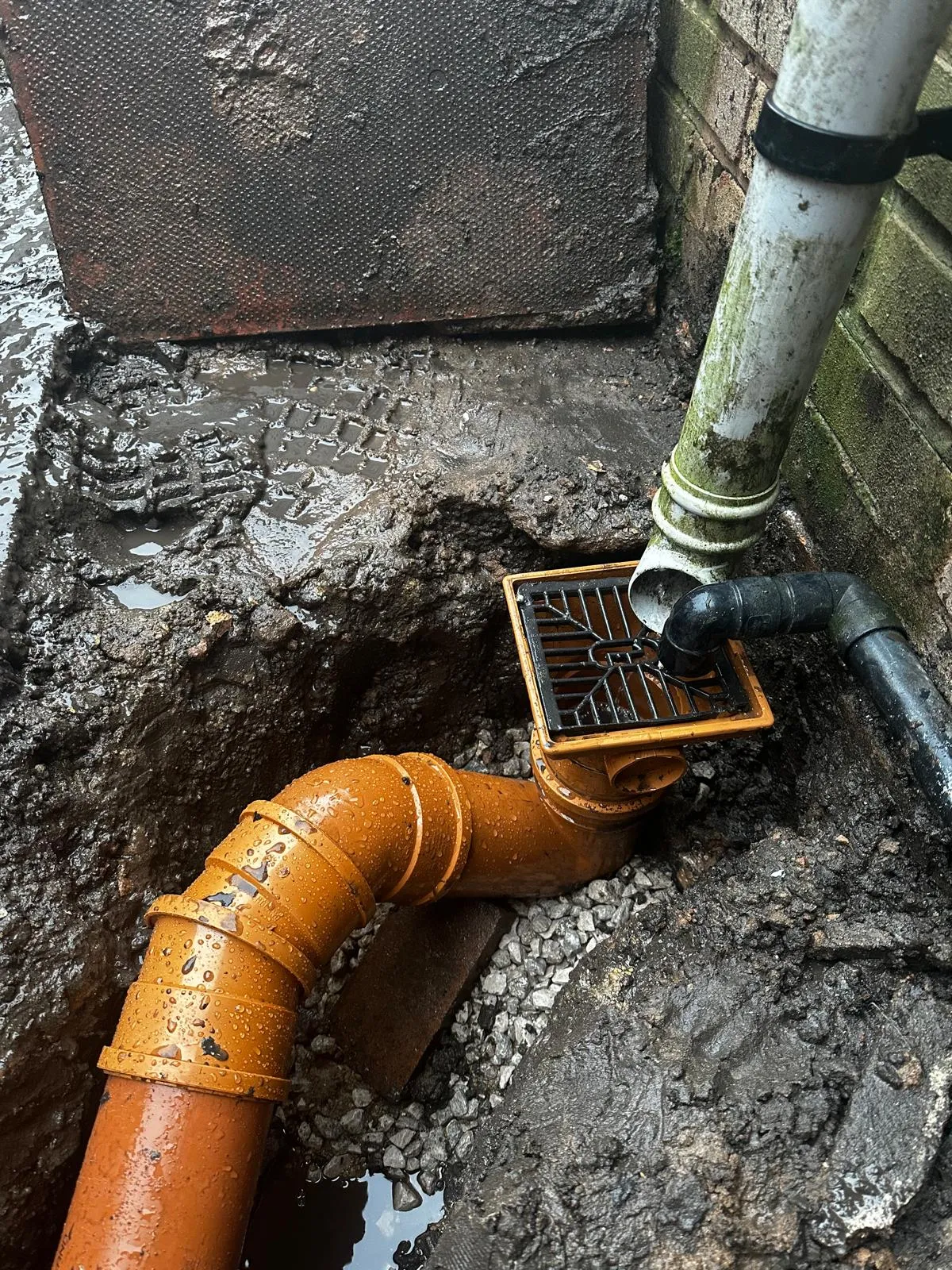 Excavated outdoor drainage system showing a gully, downpipe, and new orange pipes in a muddy trench.