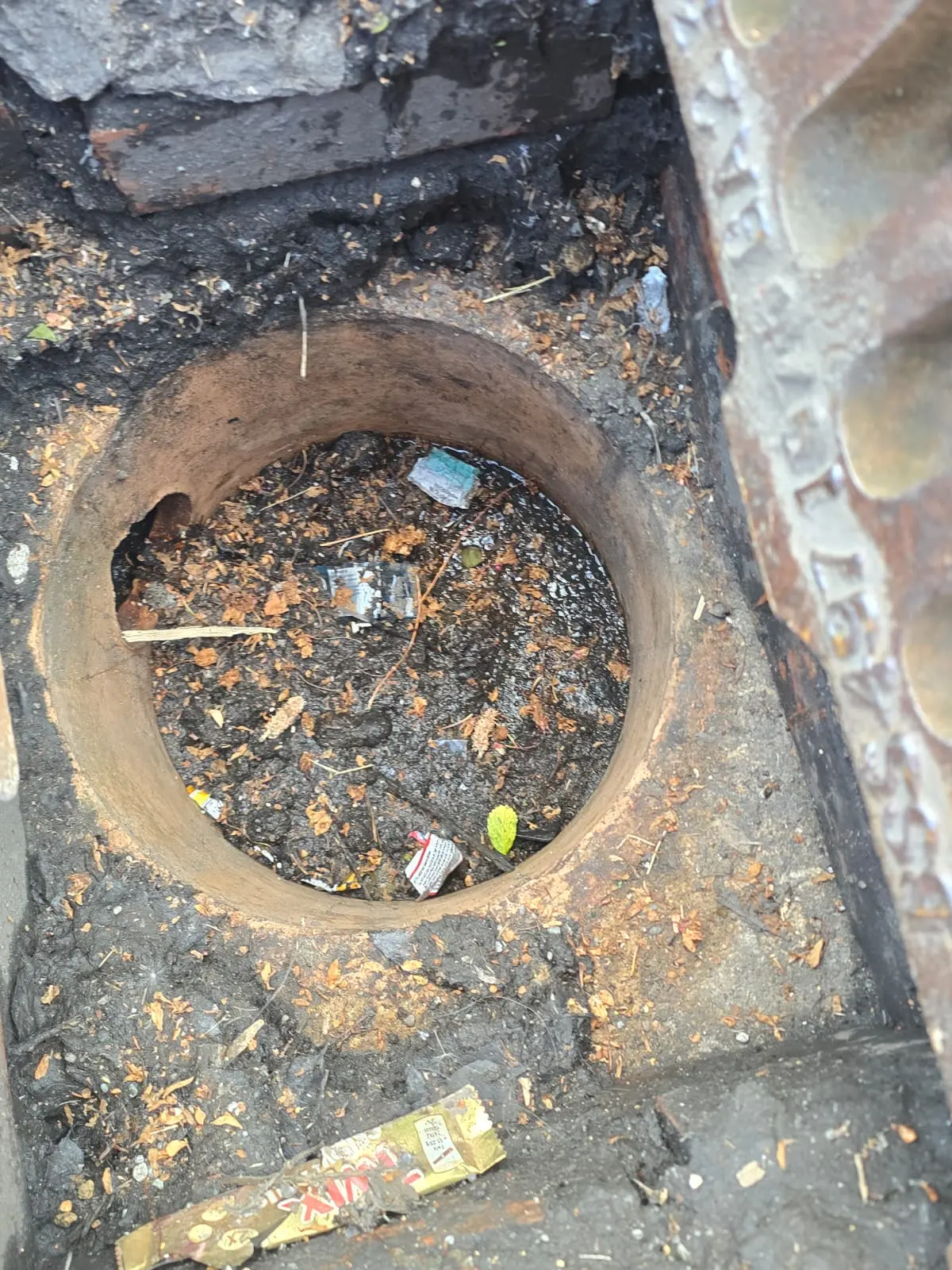 Top-down view of an open, blocked outdoor drain inspection chamber filled with dirt and debris