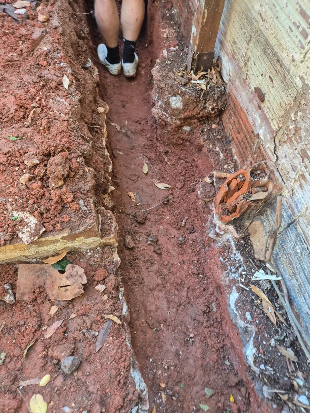 A construction worker standing in a narrow trench during groundworks next to an existing brick foundation.