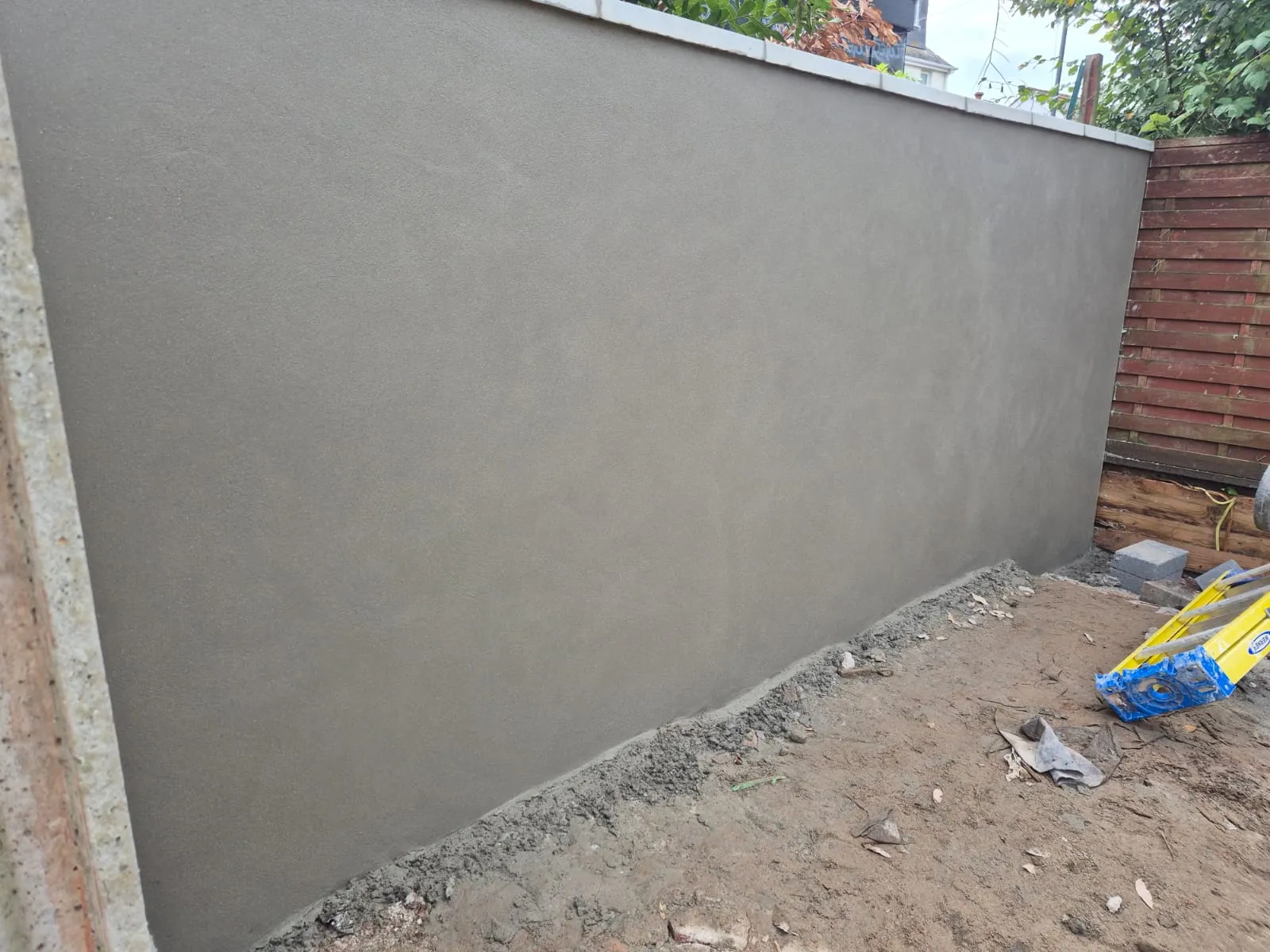 A freshly rendered grey outdoor wall, possibly a garden boundary, with new plasterwork visible and groundworks in progress.