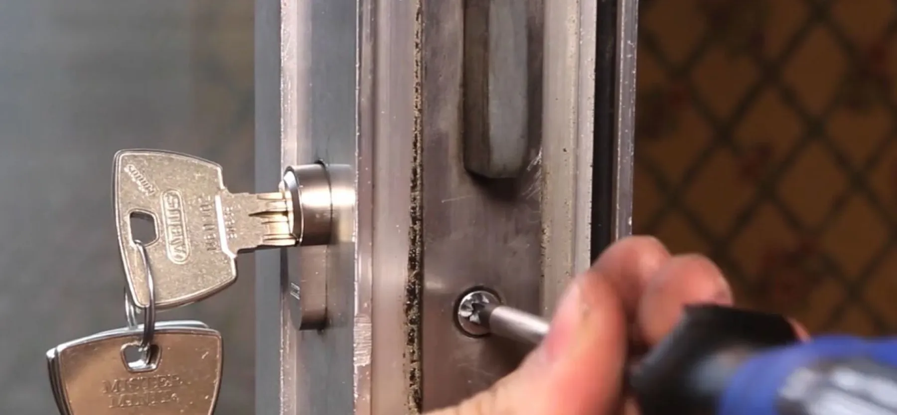 Close-up of a locksmith's hand using a screwdriver to remove a door lock cylinder with a key inserted.