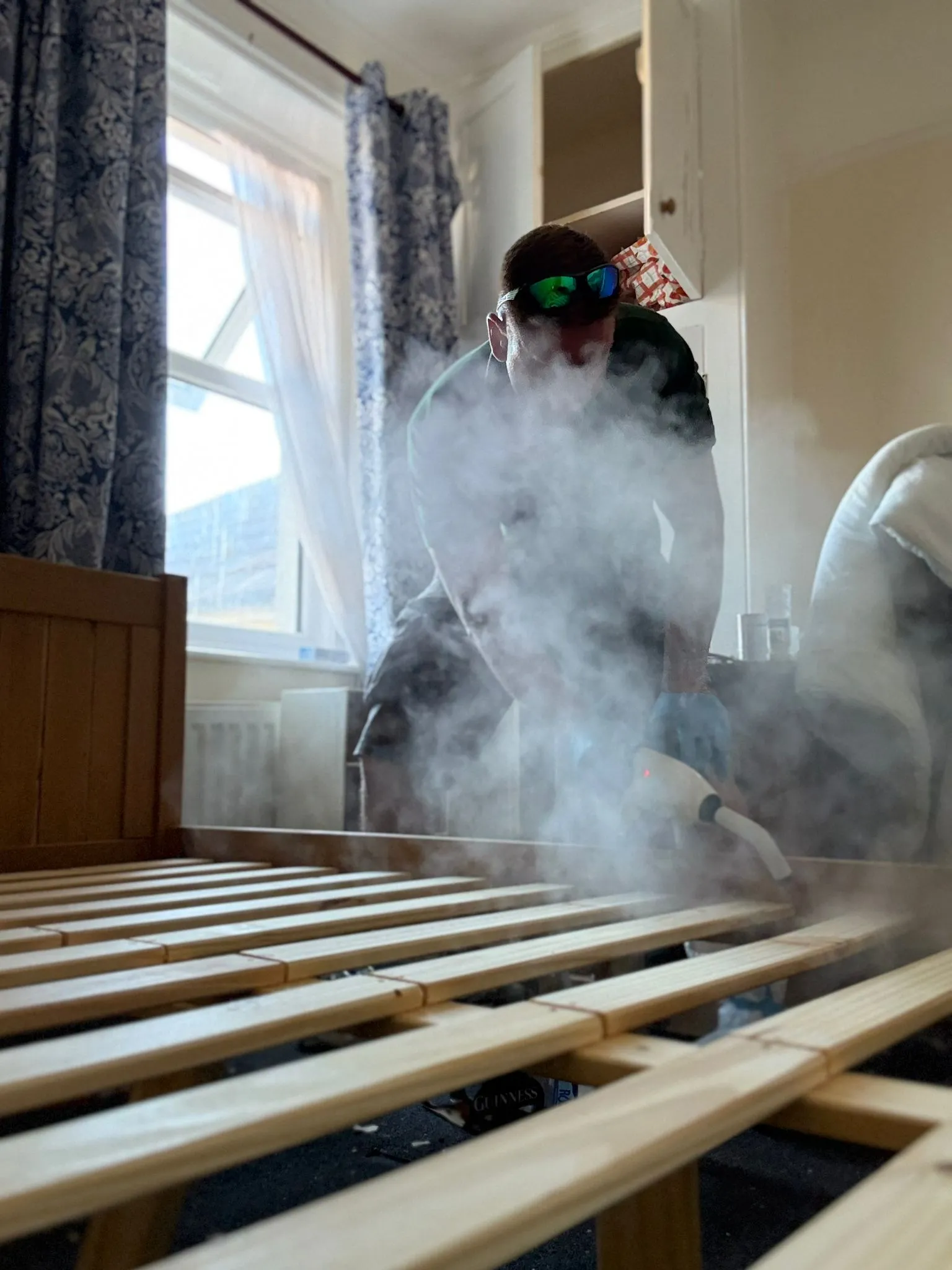A pest control technician using a steam machine to treat a wooden bed frame for bed bugs, with visible steam.