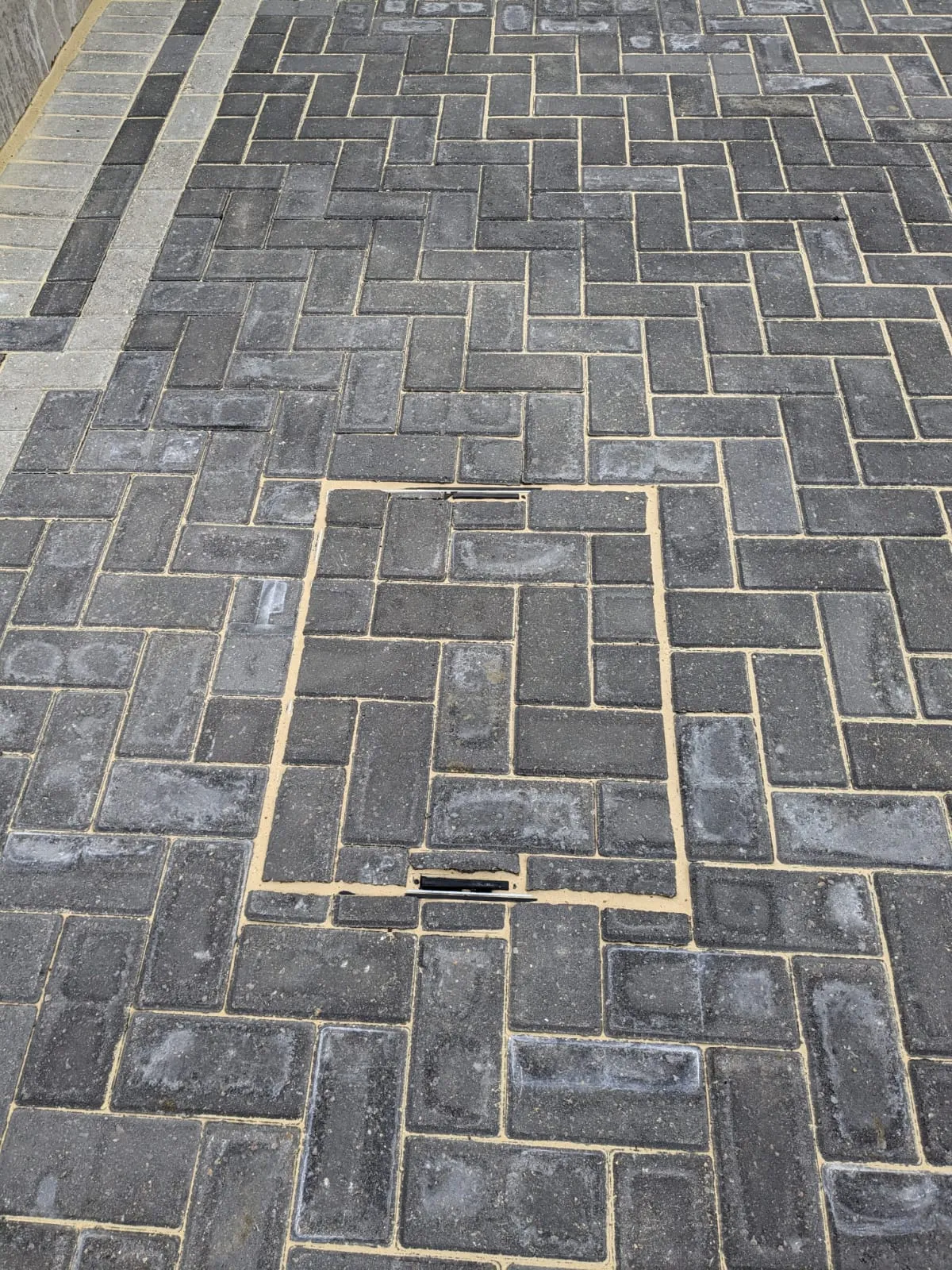 Newly installed dark grey block paving driveway with an integrated drainage manhole cover and light grey border.
