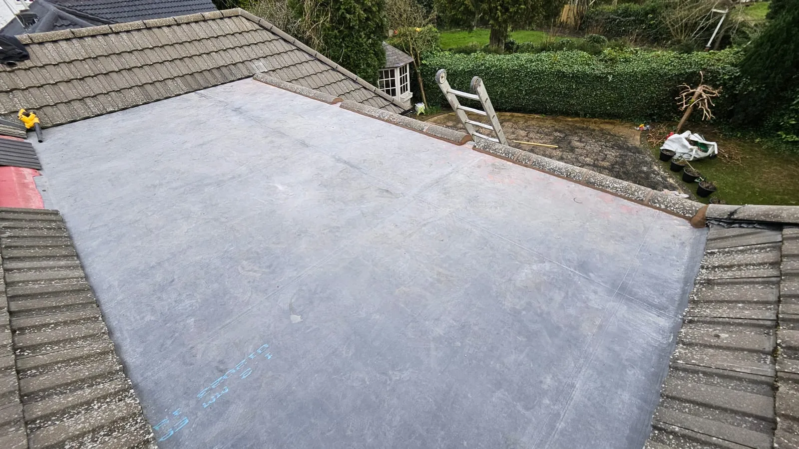 A newly installed grey flat roof membrane, possibly EPDM or felt, integrated with a tiled pitched roof section of a house, with a garden in the background.