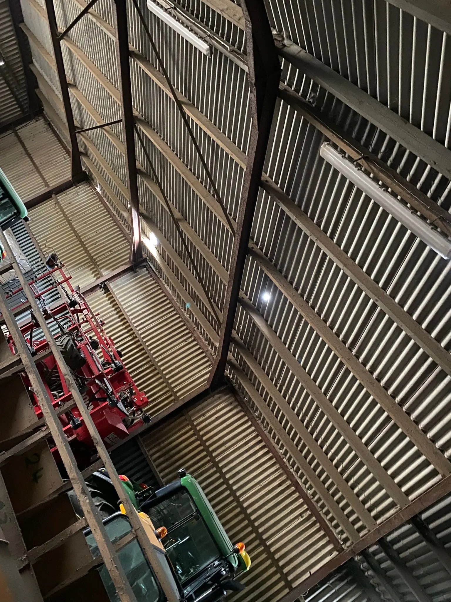 Interior of a steel-framed farm shed with corrugated roofing, housing a green tractor and other agricultural machinery.