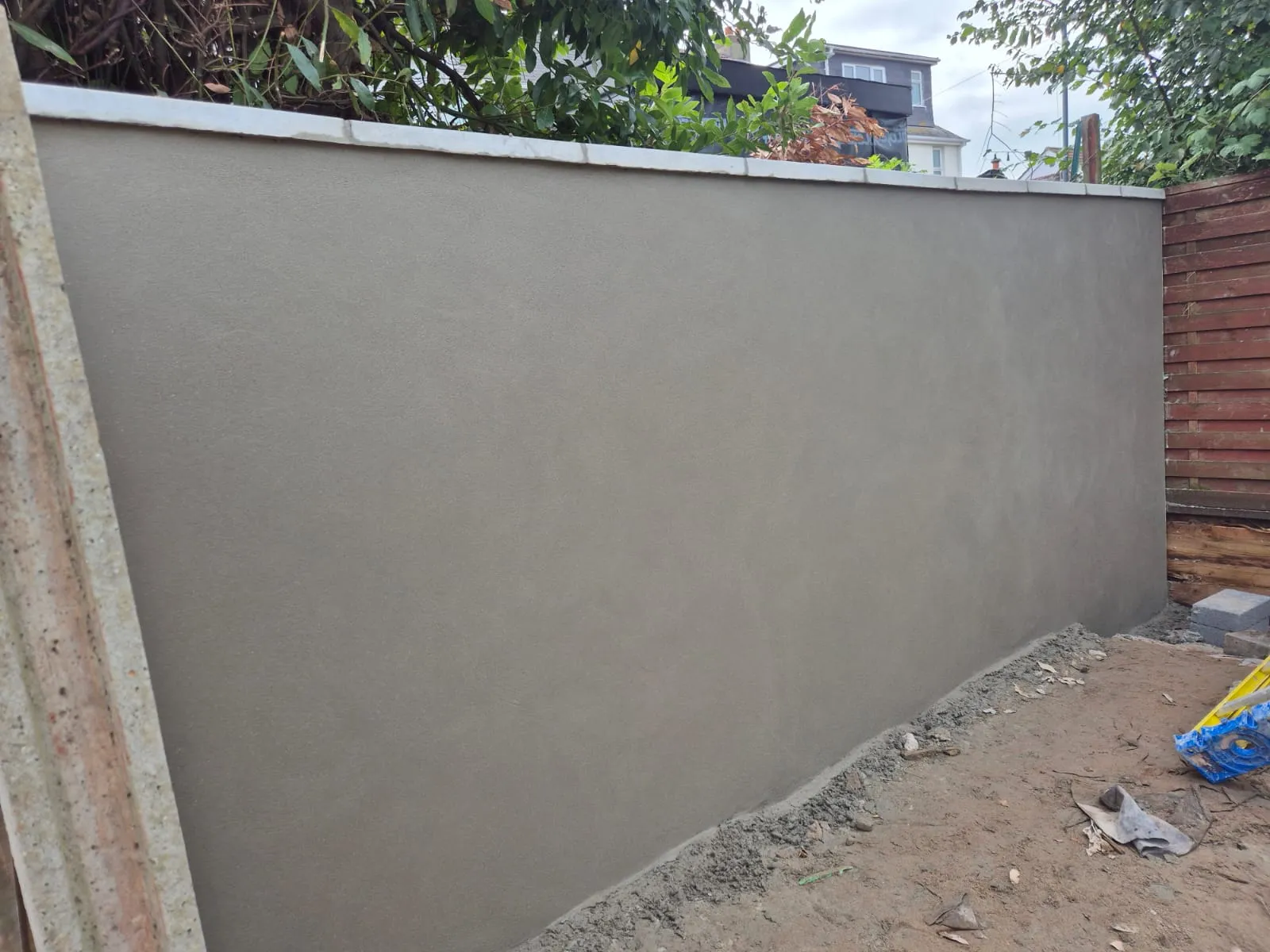 A freshly rendered grey garden wall next to a wooden fence, with some earth at its base, demonstrating quality plastering and masonry work.