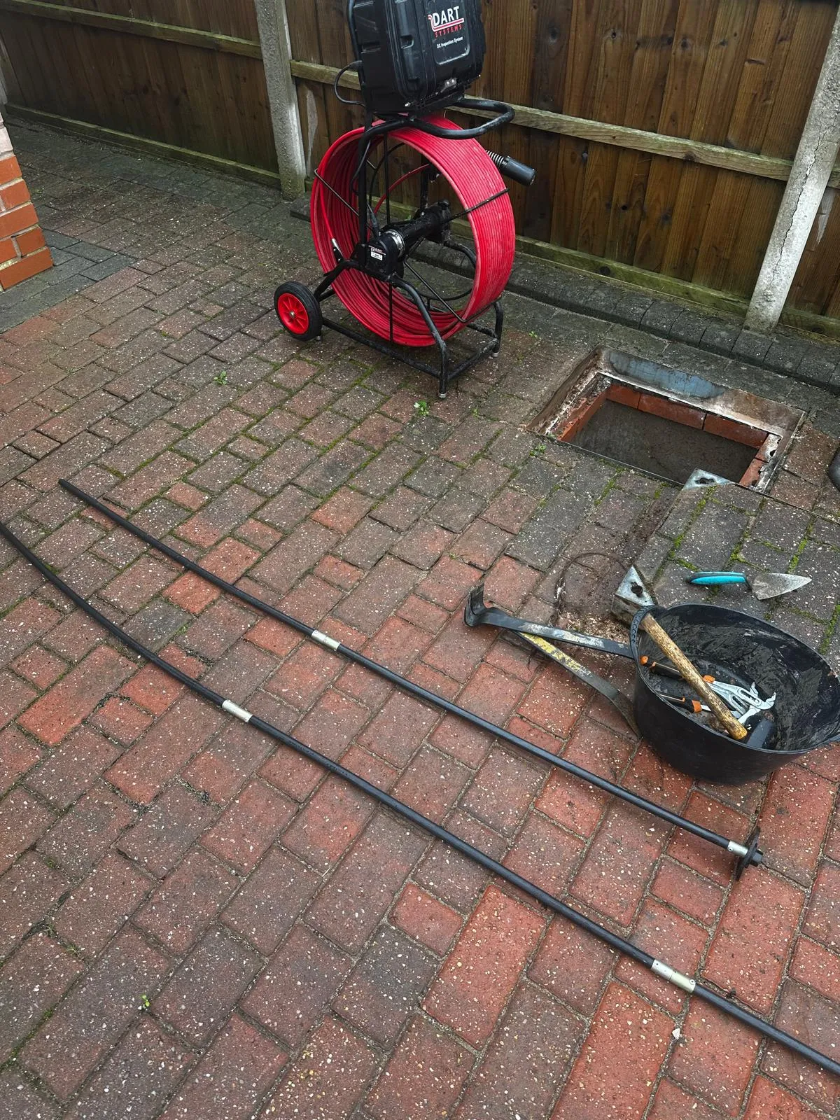 CCTV drain camera reel, drain rods, and tools next to an open manhole on a paved area, signifying a drainage survey or unblocking.