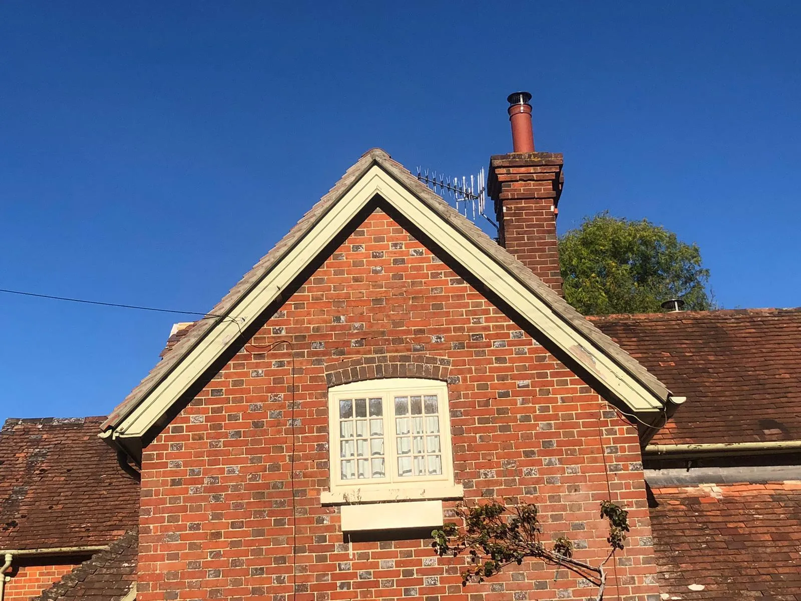 Gable end of a brick house with tiled roof, window, and chimney against a clear blue sky.