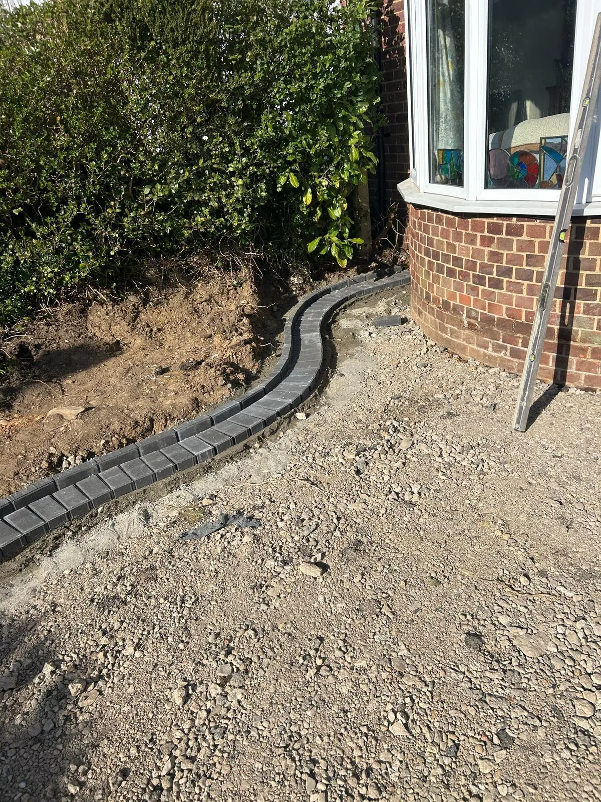 A close-up view of a new driveway under construction, featuring a neatly installed curved border of dark grey block pavers set in cement, with a layer of gravel sub-base for surfacing. A house with a brick bay window is visible in the background.