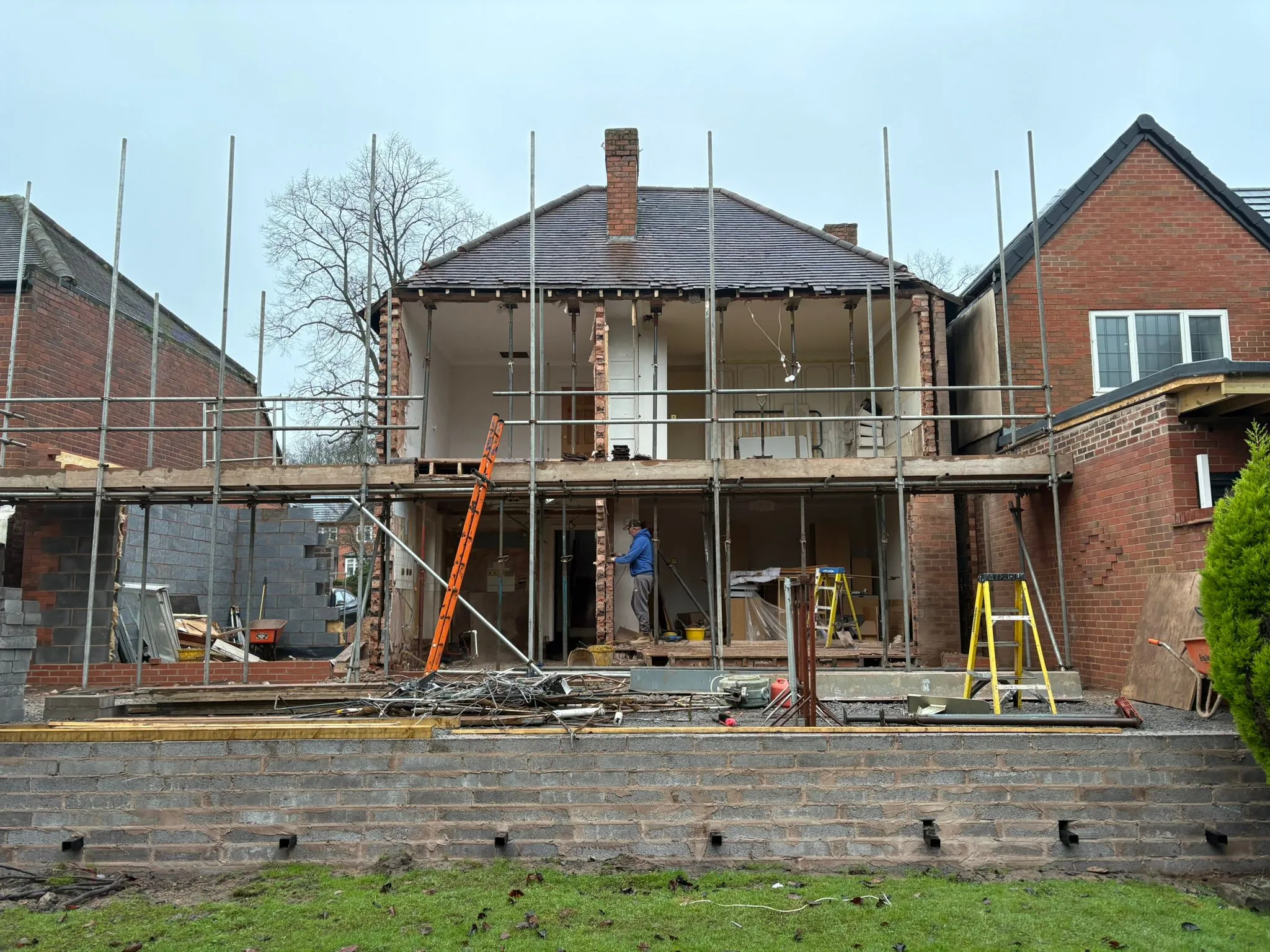 An active construction site for a home extension, showing scaffolding around the existing house structure and the new concrete block foundations for the extension. Building materials and tools are visible on the ground.