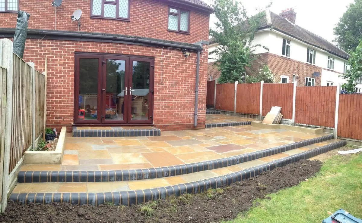 Newly laid multi-level patio with steps and paving slabs, dark brick edging, leading to a house with bi-fold doors. Fresh soil and new timber fencing are visible.