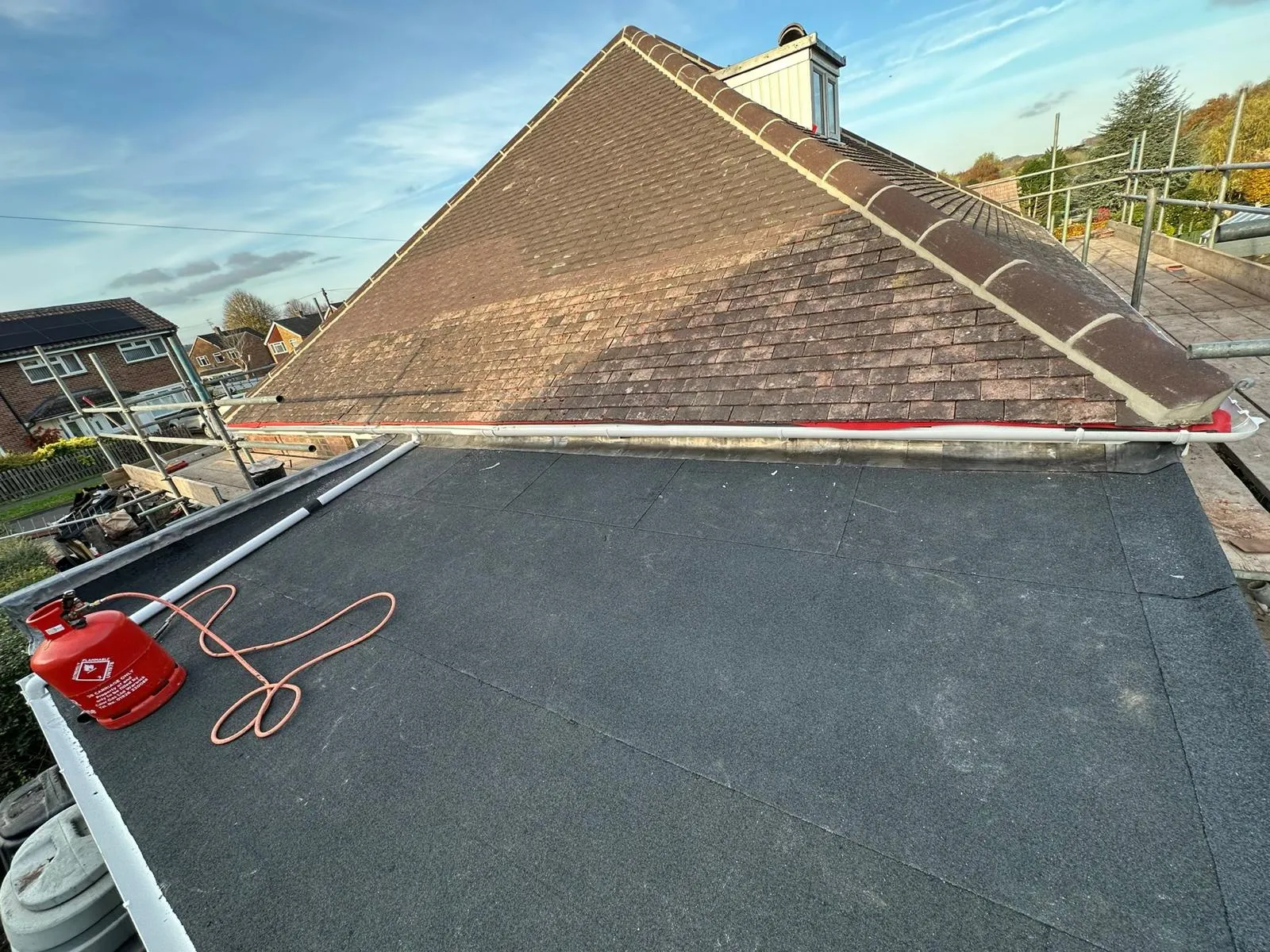 Professional installation of a new torch-on felt flat roof by Delta Roofcare Ltd, featuring a gas bottle, alongside an existing pitched tile roof and scaffolding.
