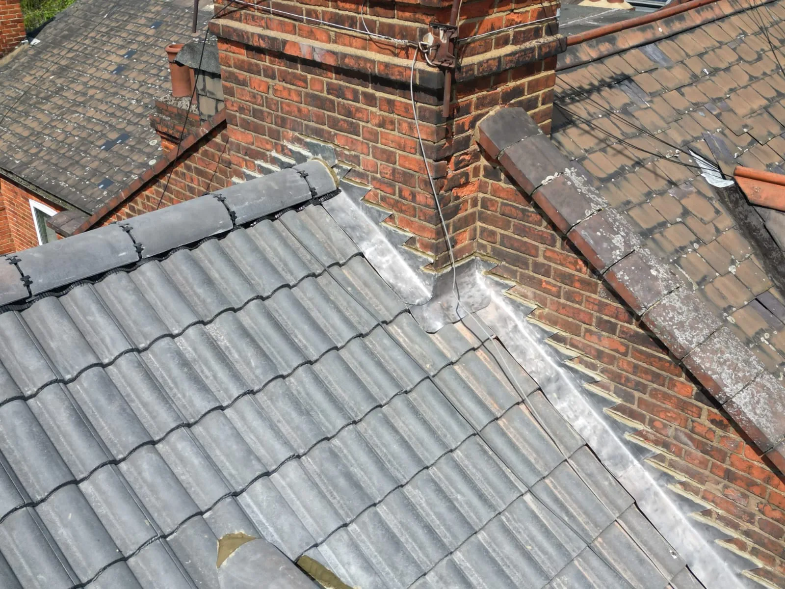 A close-up view of a residential roof showing new grey tiles, older brown tiles, and lead flashing around a brick chimney.