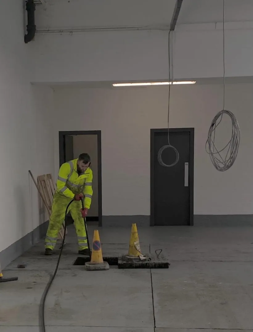 A drainage engineer using high-pressure water jetting equipment to clear a drain in what appears to be a commercial or industrial building.