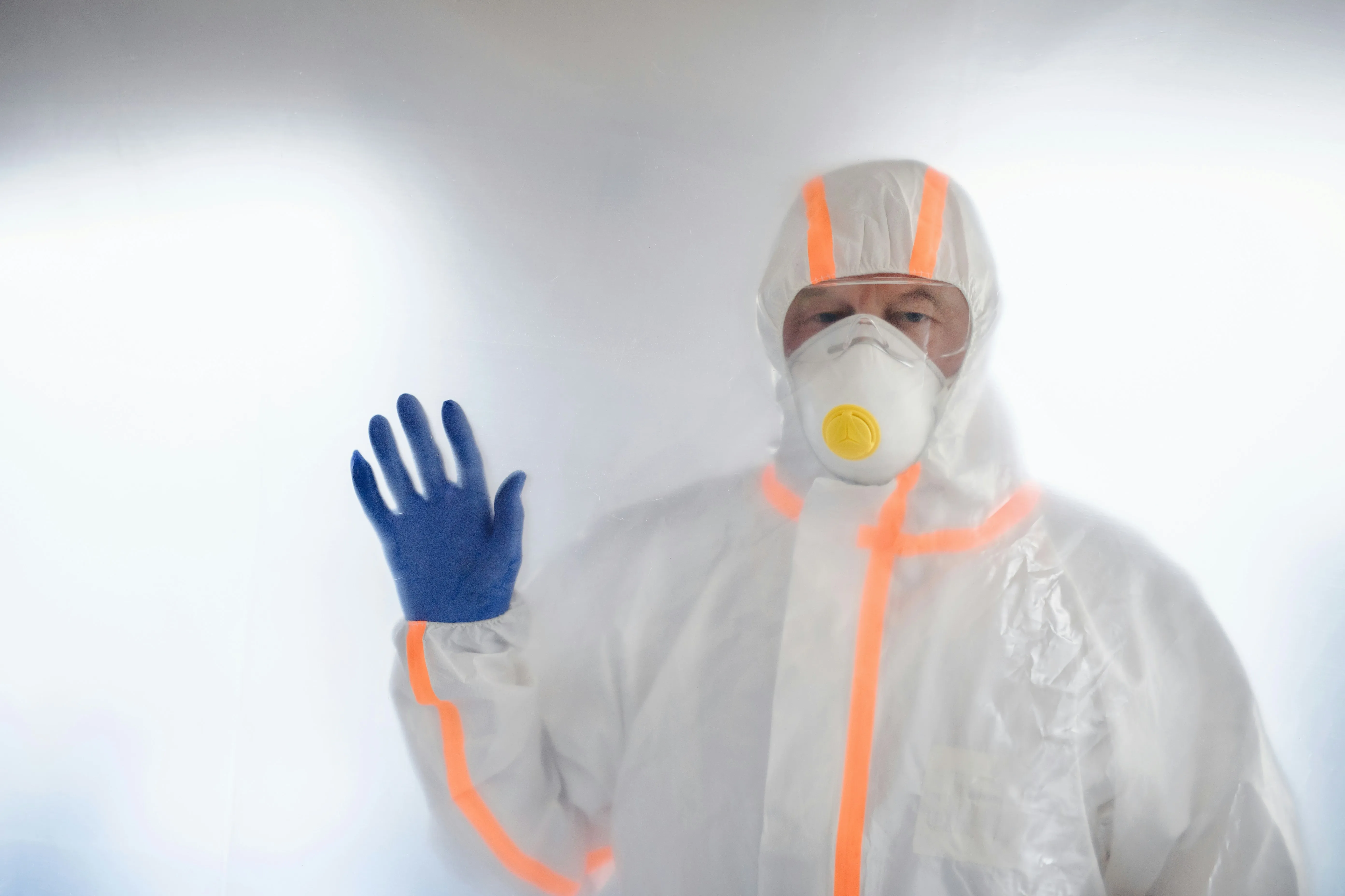 A professional wearing a white disposable hazmat suit with orange trim, a white respirator mask with a yellow filter, and blue protective gloves. Their gloved hand is pressed against a blurry, translucent barrier, symbolising safety and containment during asbestos removal work.